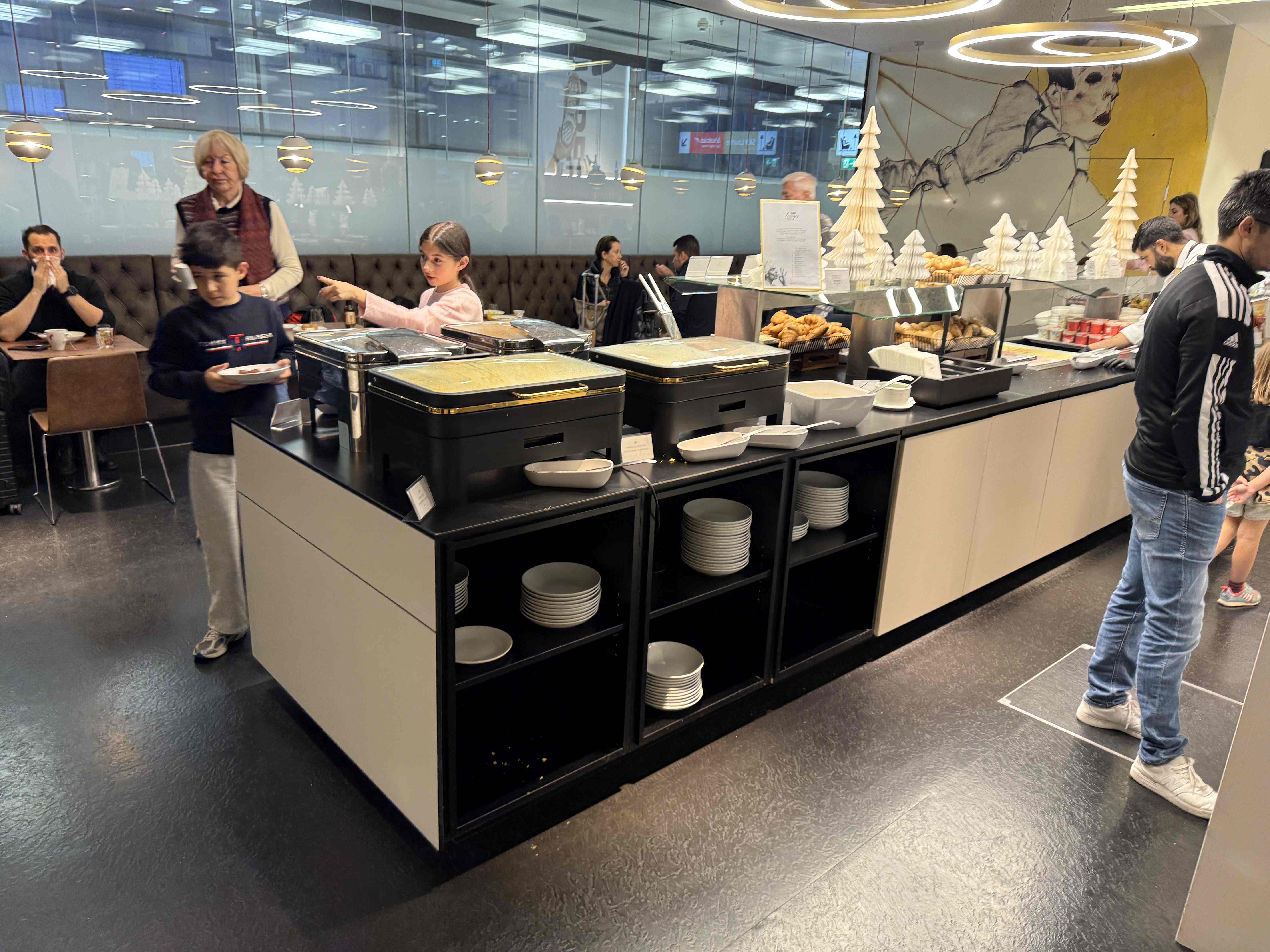 Bufet v letištní lounge: ohřívací nádoby, pečivo, talíře a cestující
