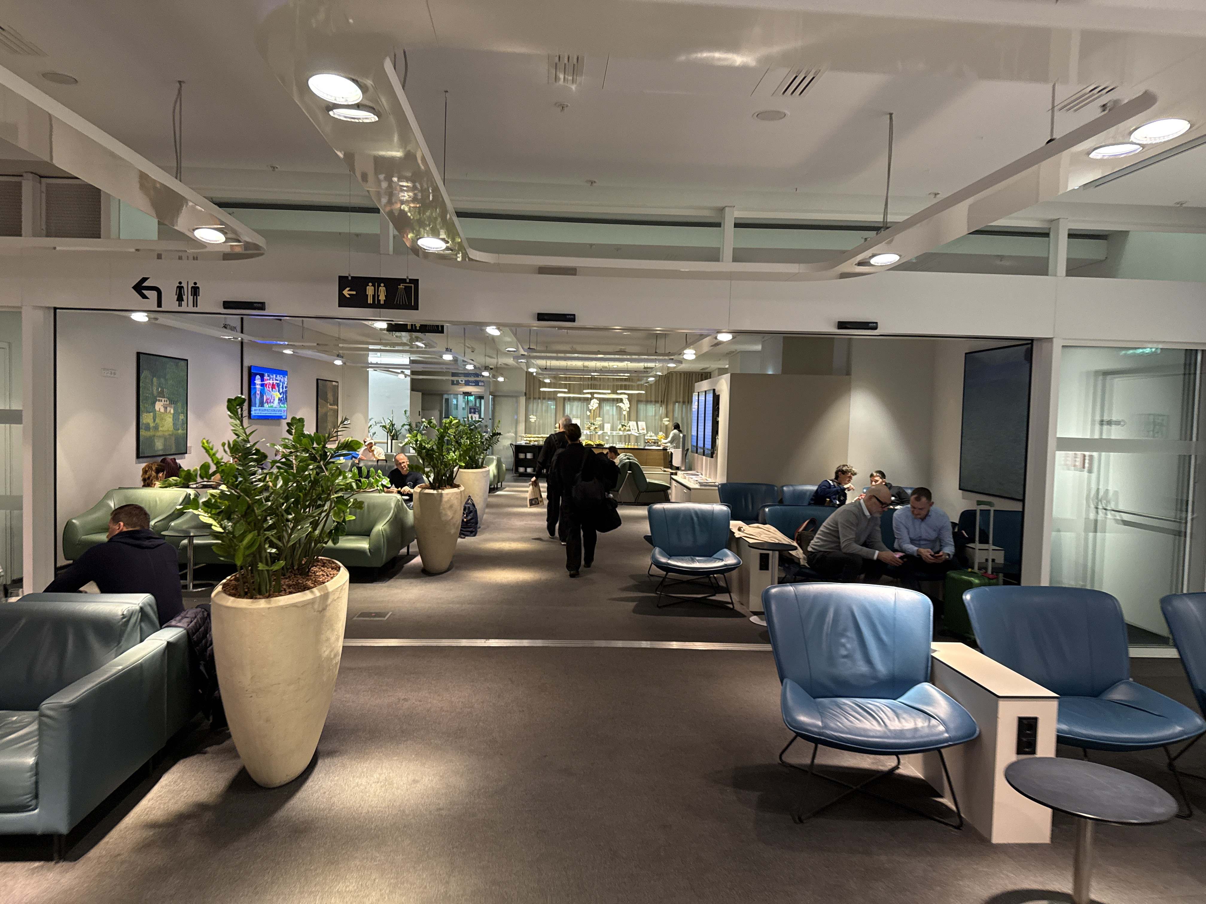 Letištní salonek s modrými křesly, zelenými rostlinami a cestujícími, směrovky k toaletám.