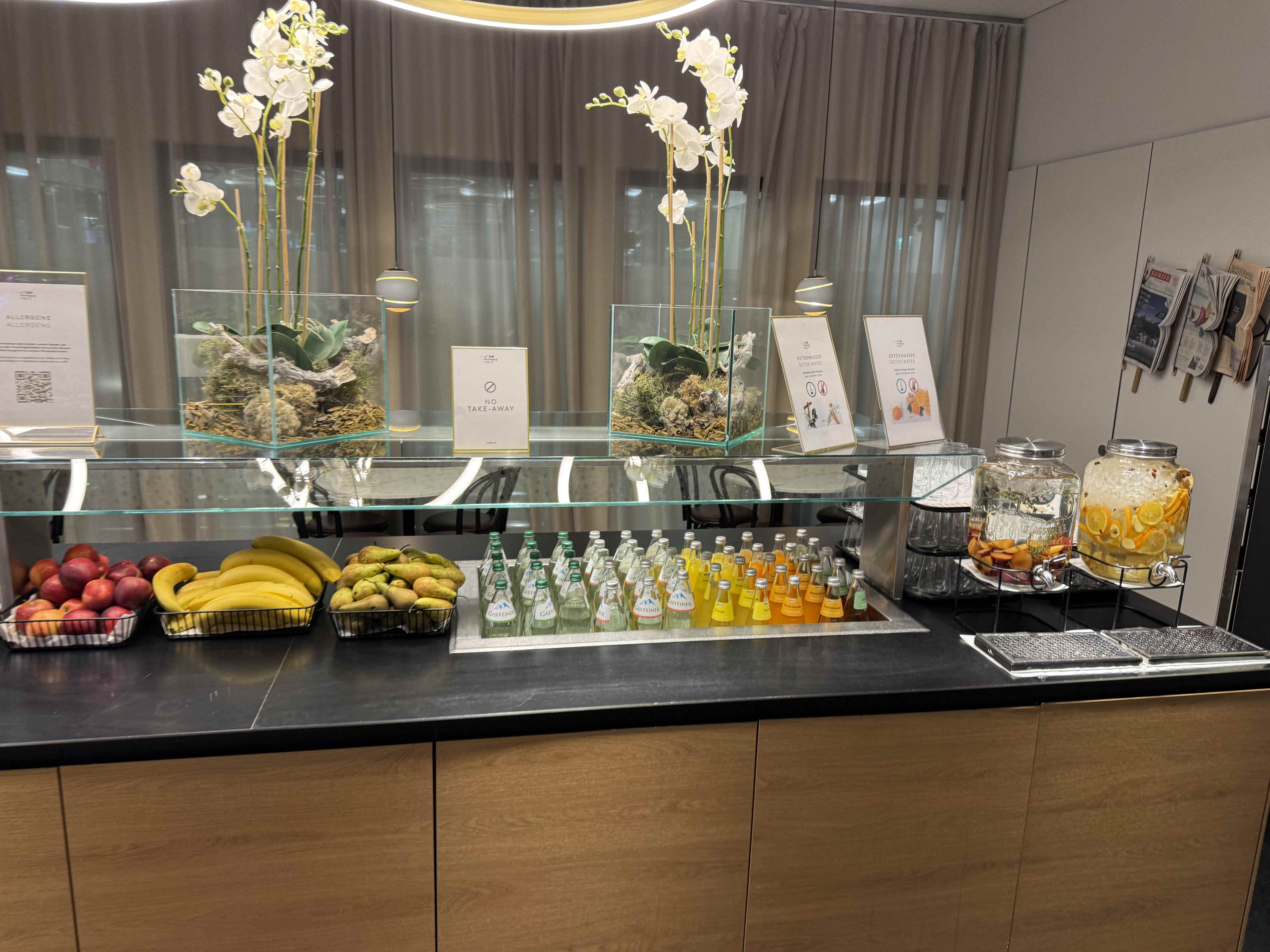 Bufet v letištním salonku: ovoce, limonády, voda, nádoby s džusem, cedule NO TAKE-AWAY.