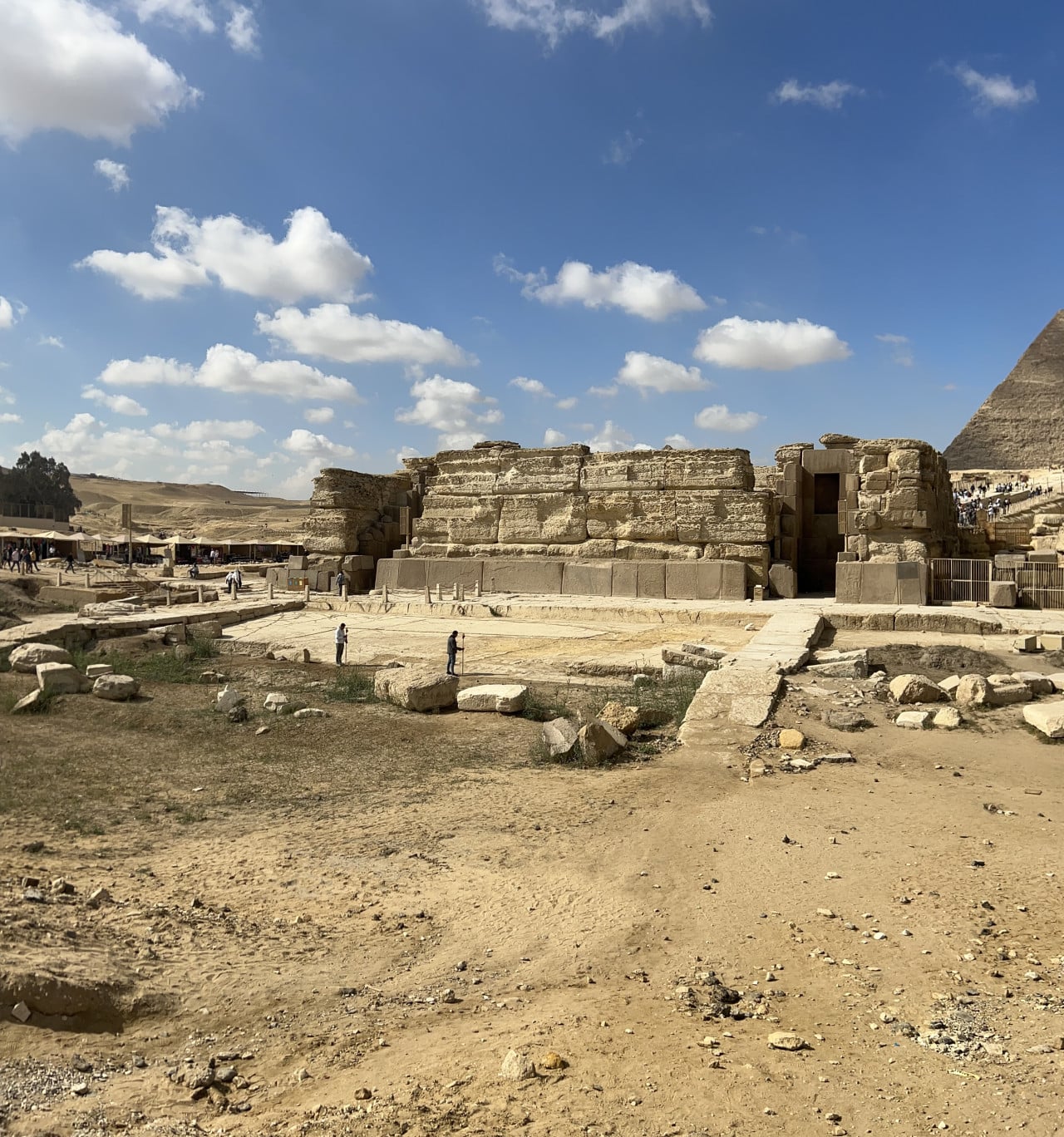 Ruiny kamenné stavby v Gíze s částí pyramidy vpravo a modrou oblohou.