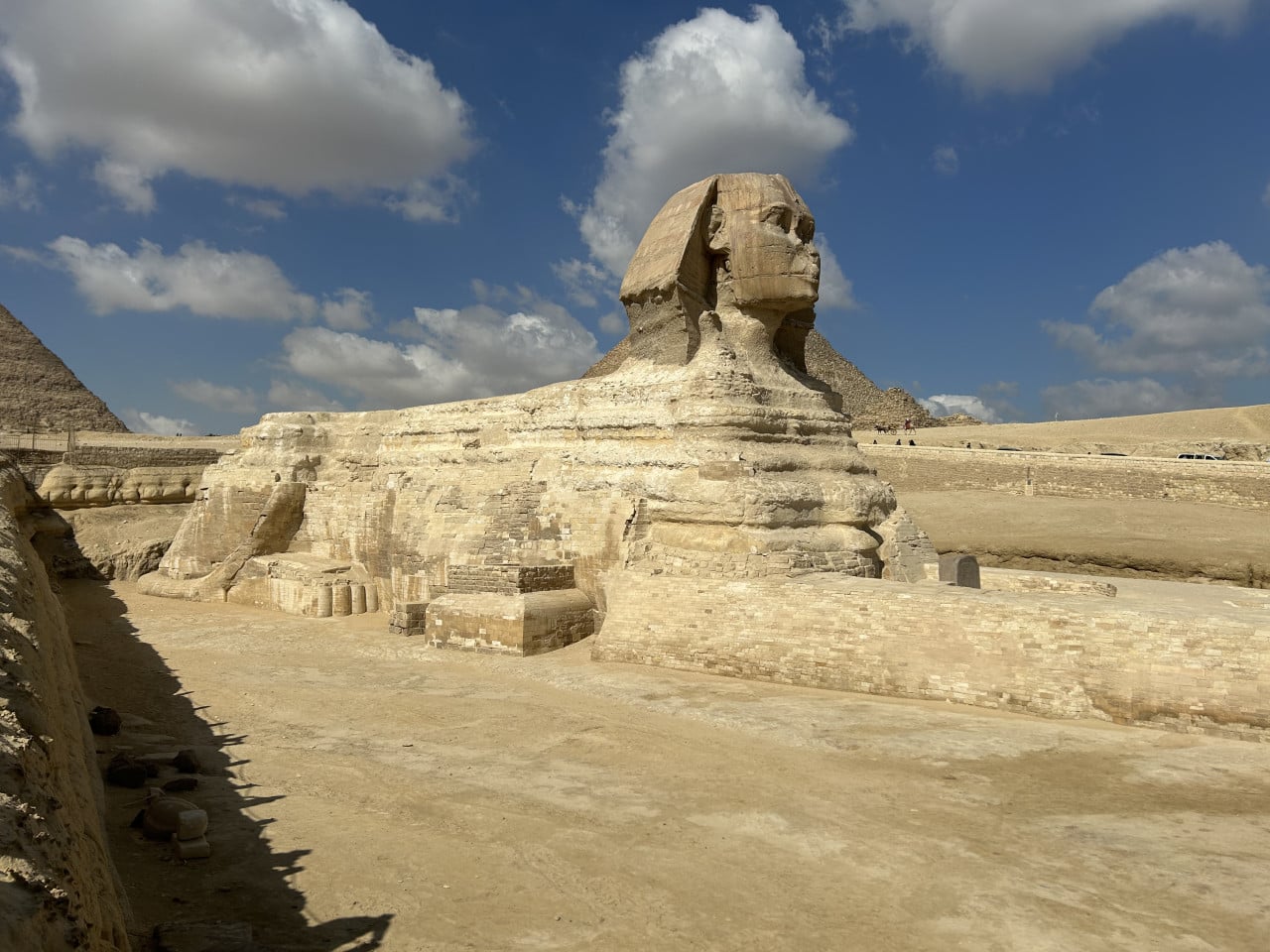 Velká sfinga v Gíze pod modrou oblohou s mraky, pyramidy v pozadí