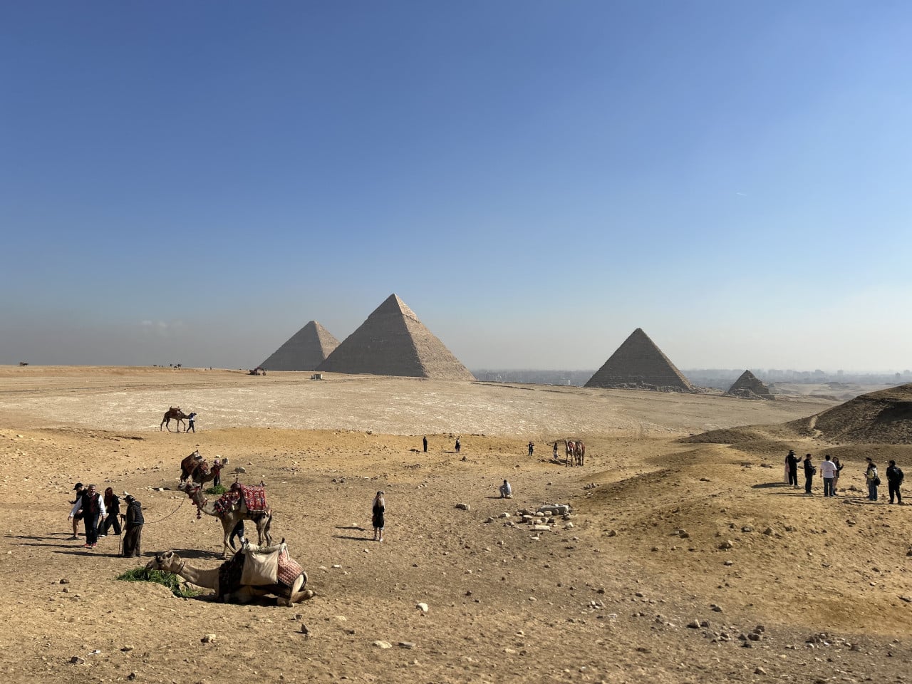 Pyramidy v Gíze, turisté a velbloudi na pouštní planině pod modrou oblohou