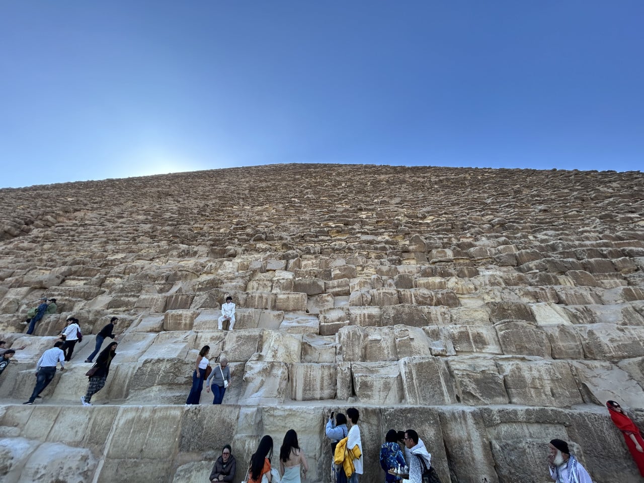 Pohled na obrovské kamenné bloky Cheopsovy pyramidy s turisty pod modrou oblohou