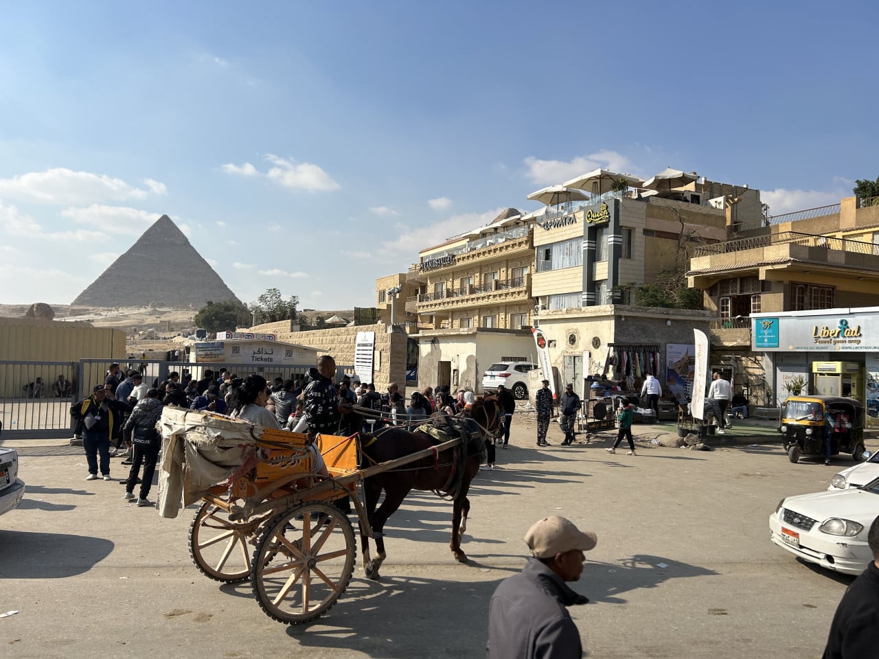 Pyramida v Gíze, turisté, koňský povoz a okolní budovy pod modrou oblohou.