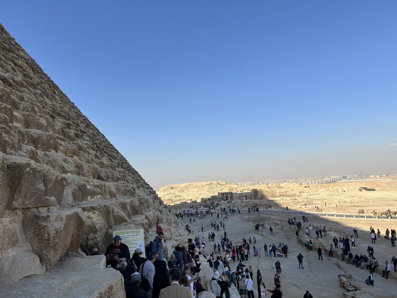 Strmý kamenný bok Cheopsovy pyramidy s davem turistů a pouštní krajinou v Gíze