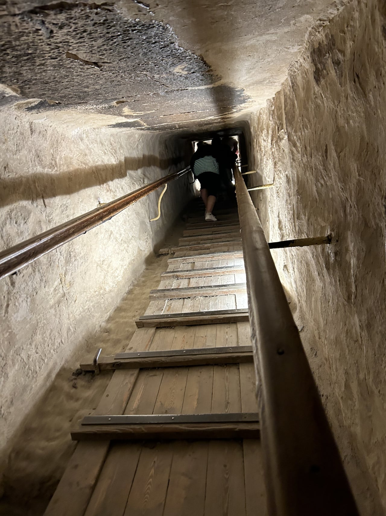 Úzká kamenná chodba uvnitř pyramidy s dřevěnými schody a zábradlím, lidé stoupají vzhůru.
