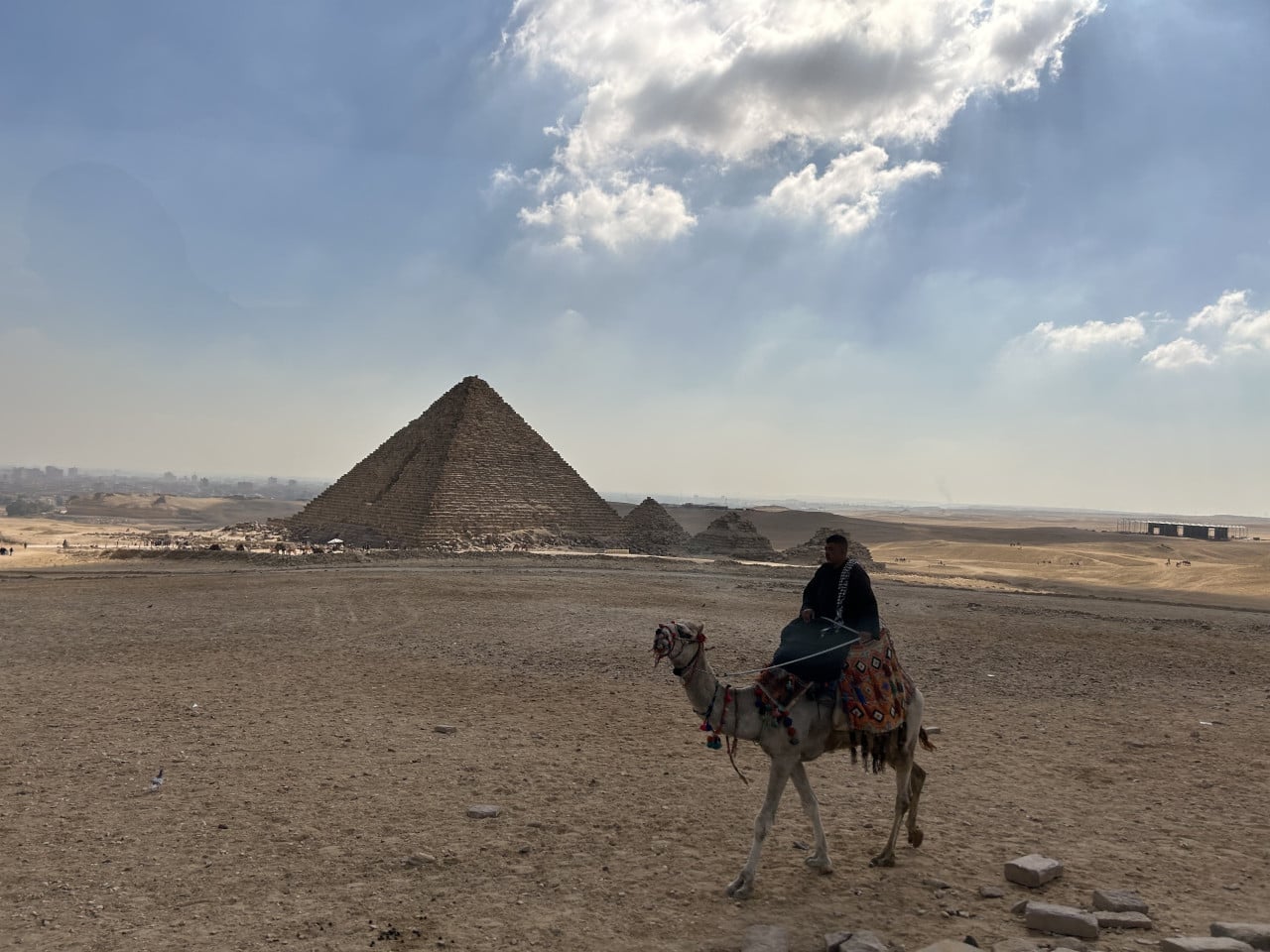 Velká pyramida v Gíze, jezdec na velbloudu v popředí, poušť a obloha s mraky.