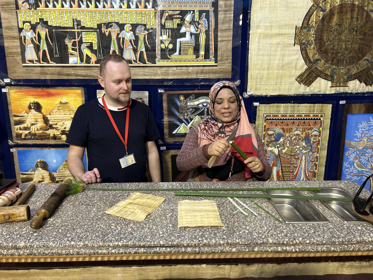 Dva lidé ukazují výrobu papyrusu před egyptskými malbami a Gízou.