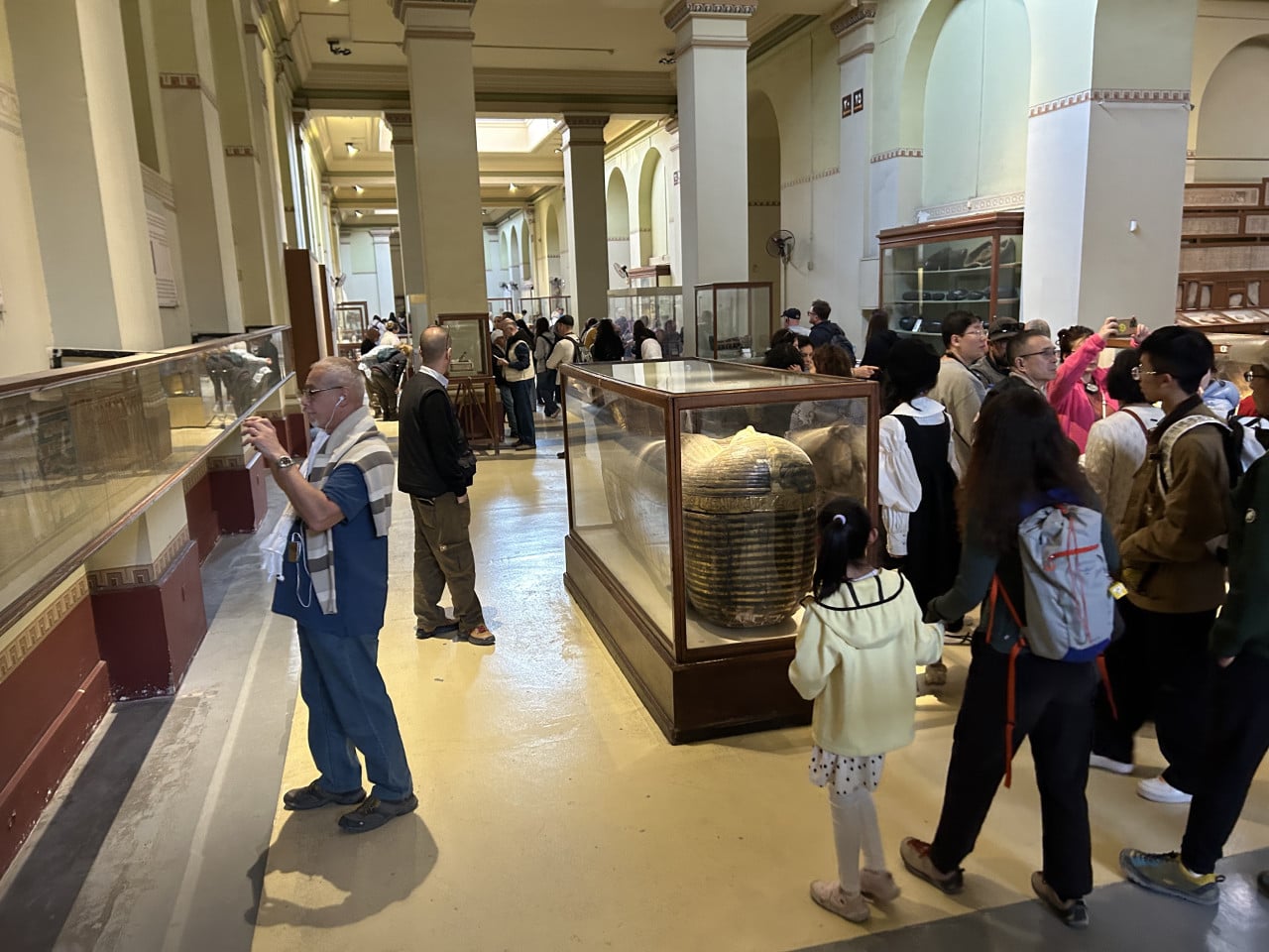 Návštěvníci v egyptském muzeu obdivují sarkofágy a exponáty ve vitrínách.