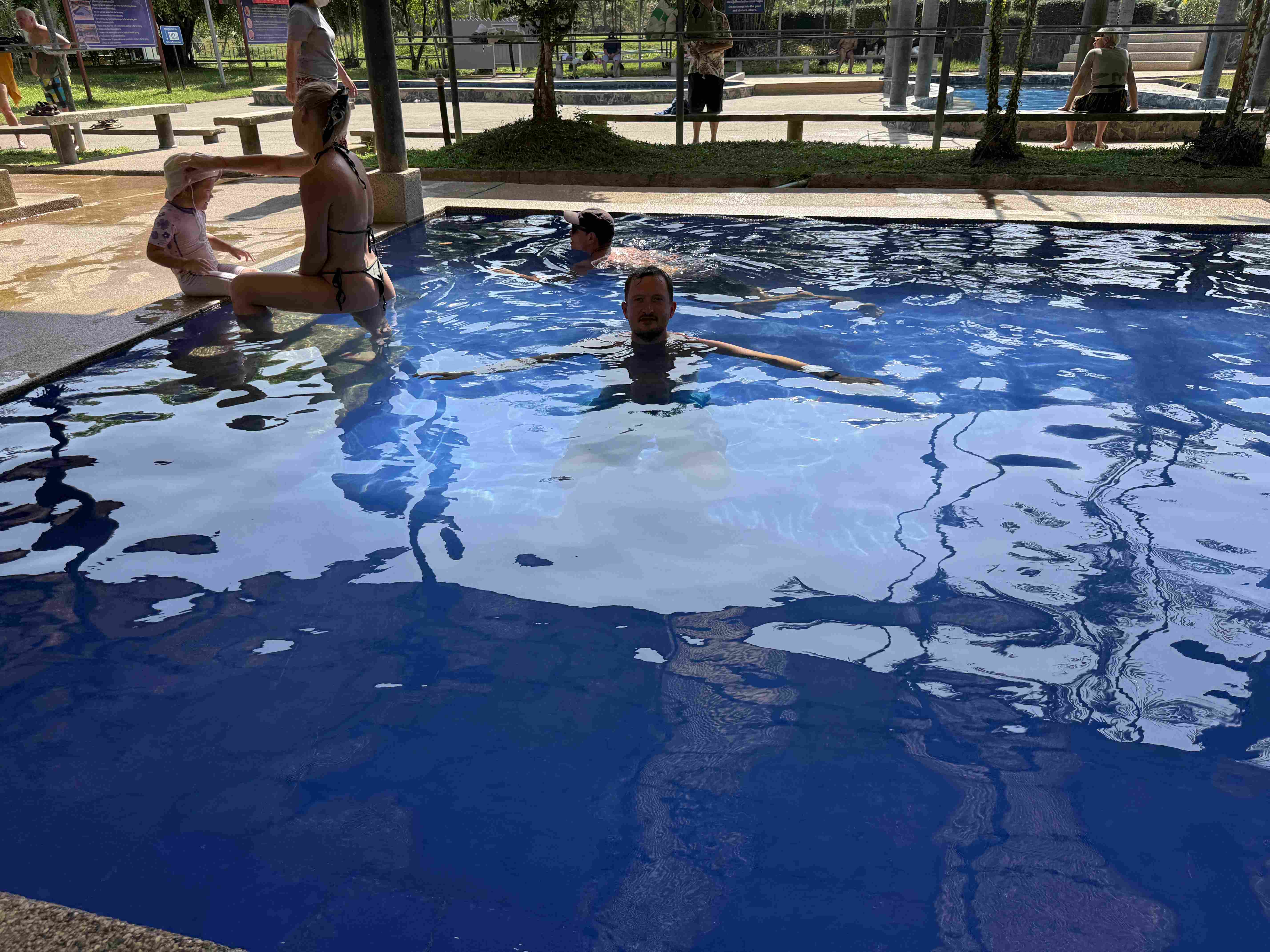 Muž plave v modrém bazénu, žena s dítětem sedí na okraji.