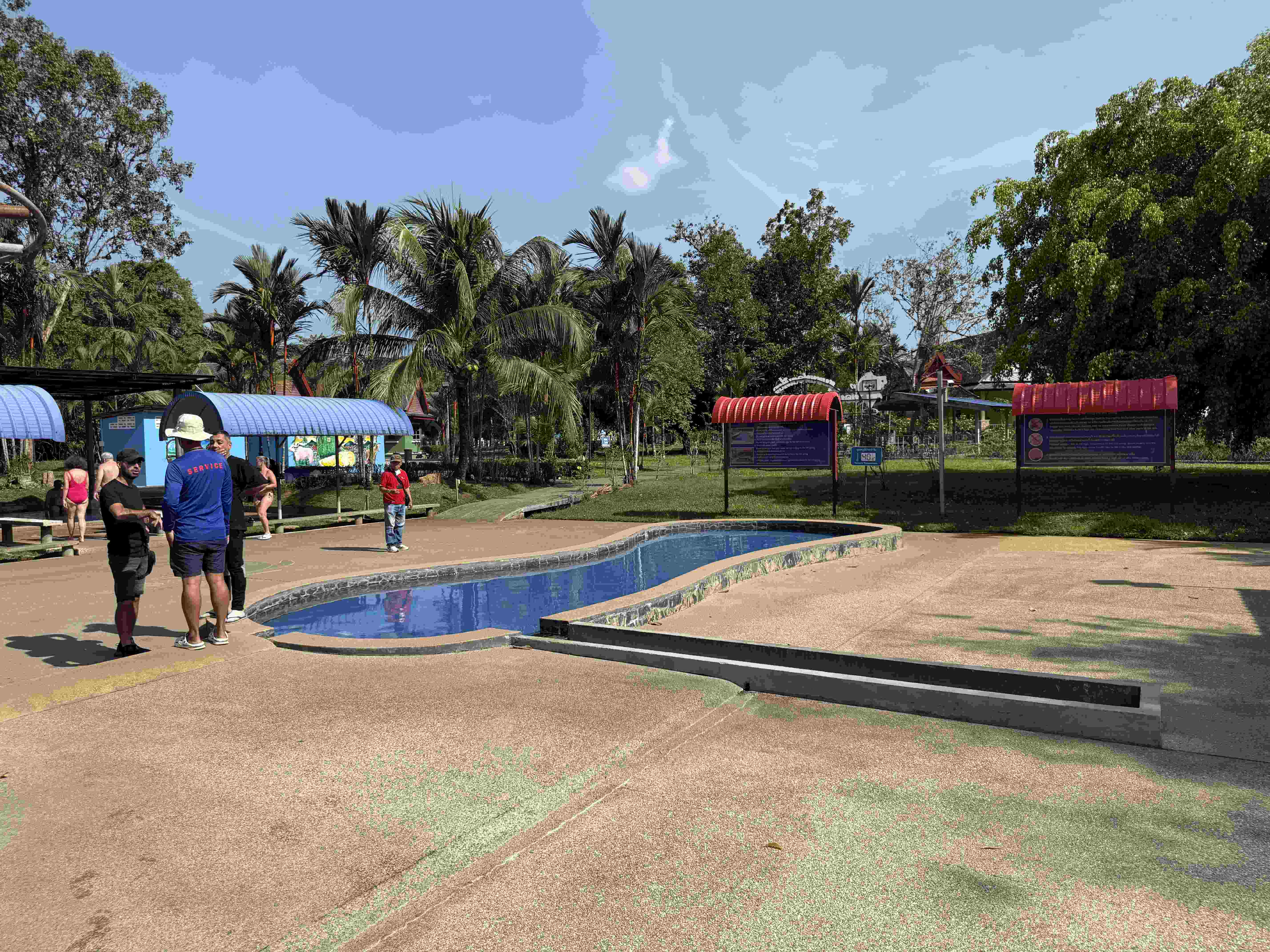 Úzký bazének v areálu Rommanee Hot Spring, lidé, palmy a informační tabule