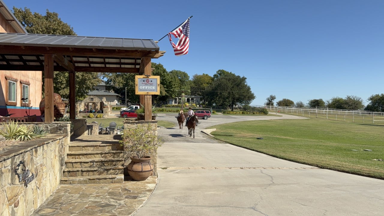 Vjezd na Buffalo Ranch s americkou vlajkou, nápisem Office a jezdci na koních.
