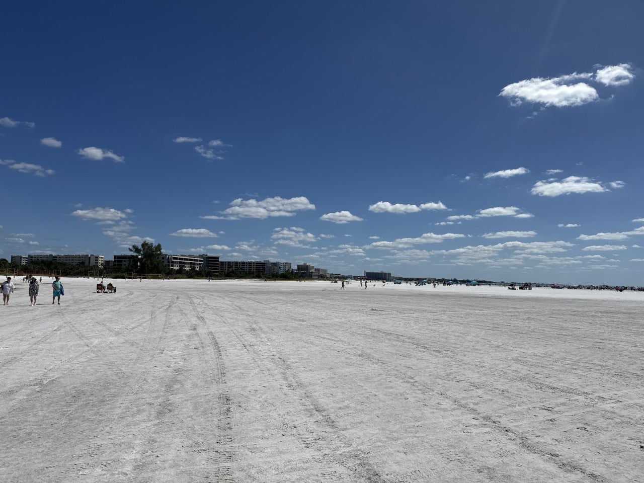 Široká bílá pláž Siesta Key s lidmi, budovami v dálce a modrou oblohou s mraky