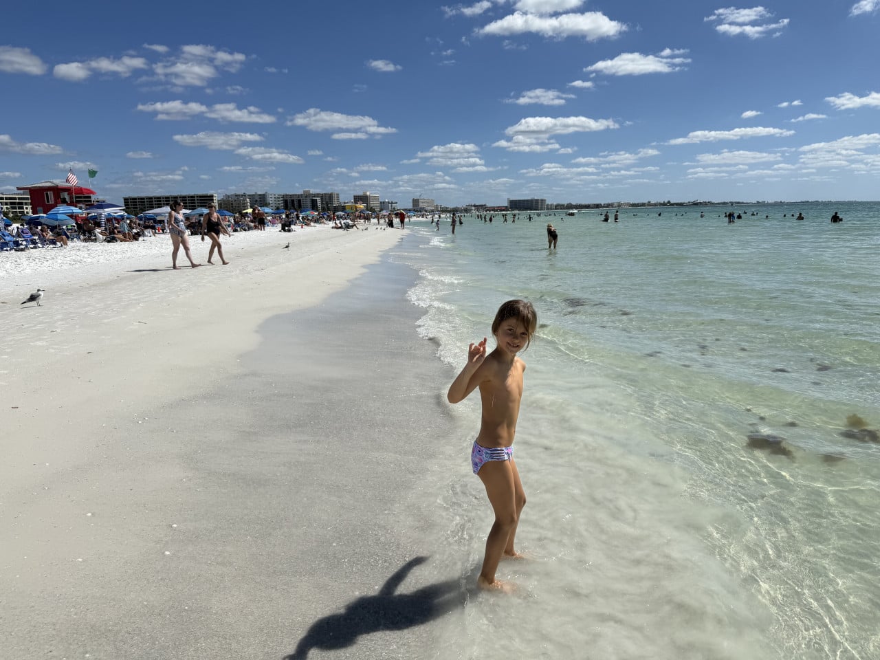 Dítě stojí u vody na Siesta Key Beach, v pozadí pláž s lidmi a slunečníky.