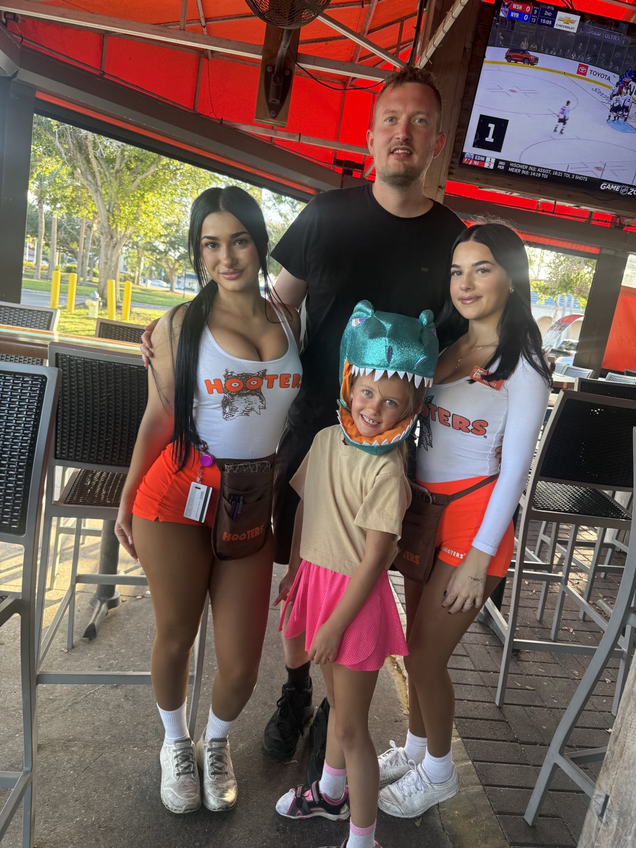 Muž a dítě s dvěma ženami v Hooters, v pozadí televize s hokejem.