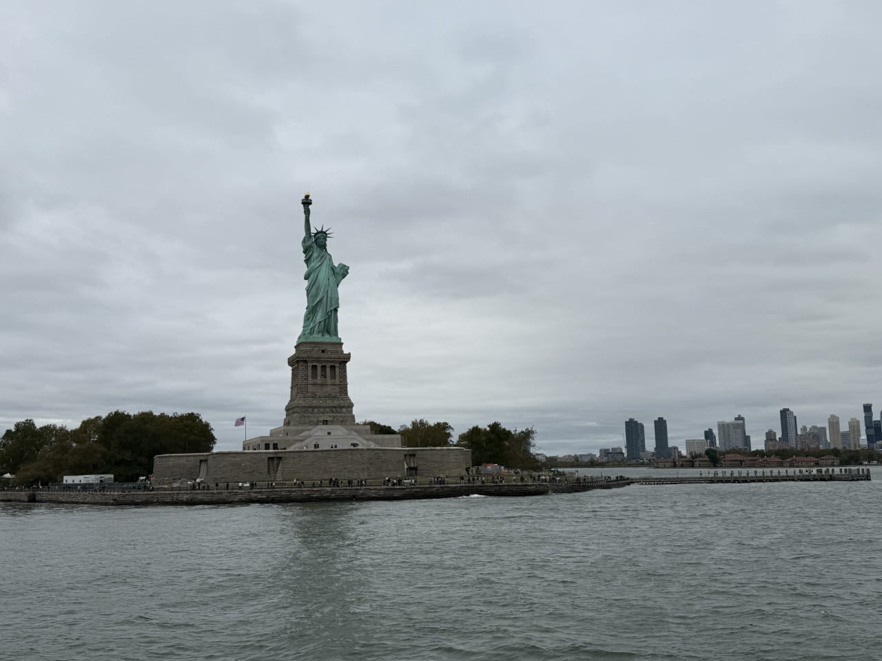 Socha Svobody na Liberty Islandu s manhattanským skyline v pozadí תחת cloudy sky