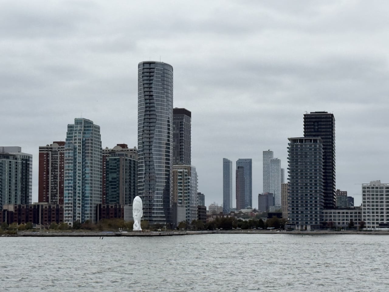 Bílá obří hlava sochy u řeky před newyorskými mrakodrapy a šedou oblohou