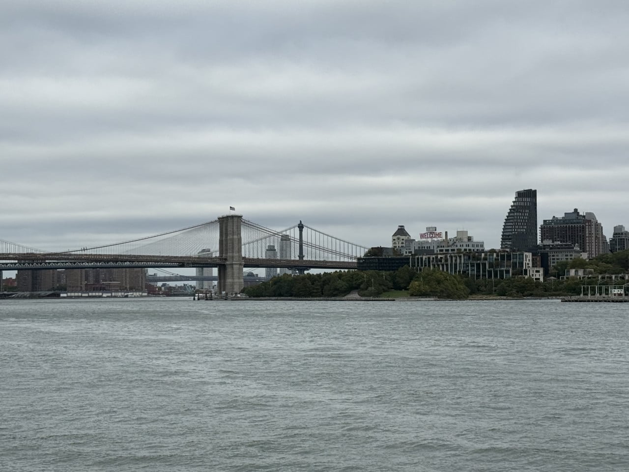 Brooklyn Bridge nad řekou, panorama Manhattanu a nápis WELCOME na budově.