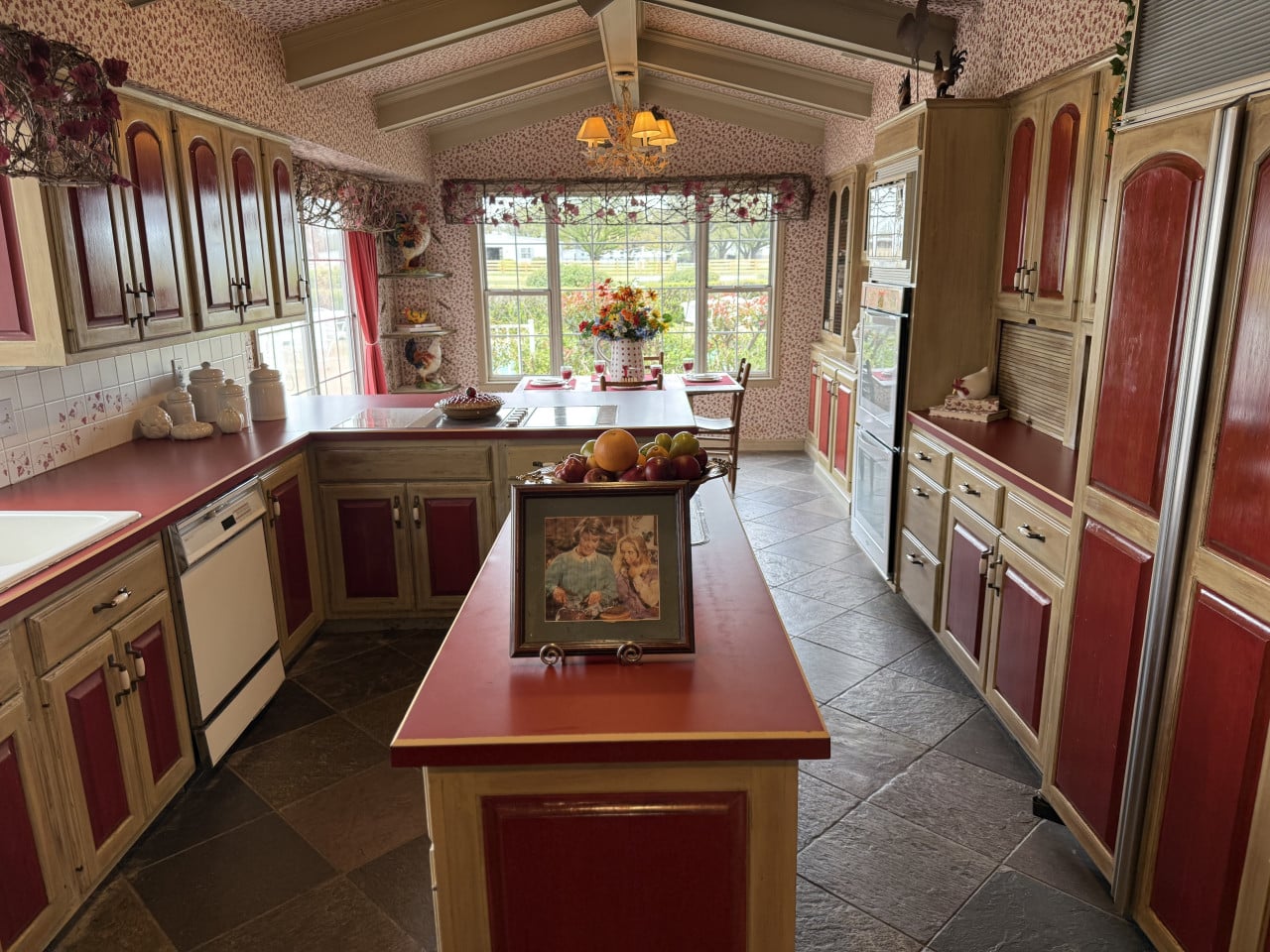 Interiér kuchyně Southfork Ranch s červenými skříňkami, ostrůvkem, ovocem a velkým oknem.