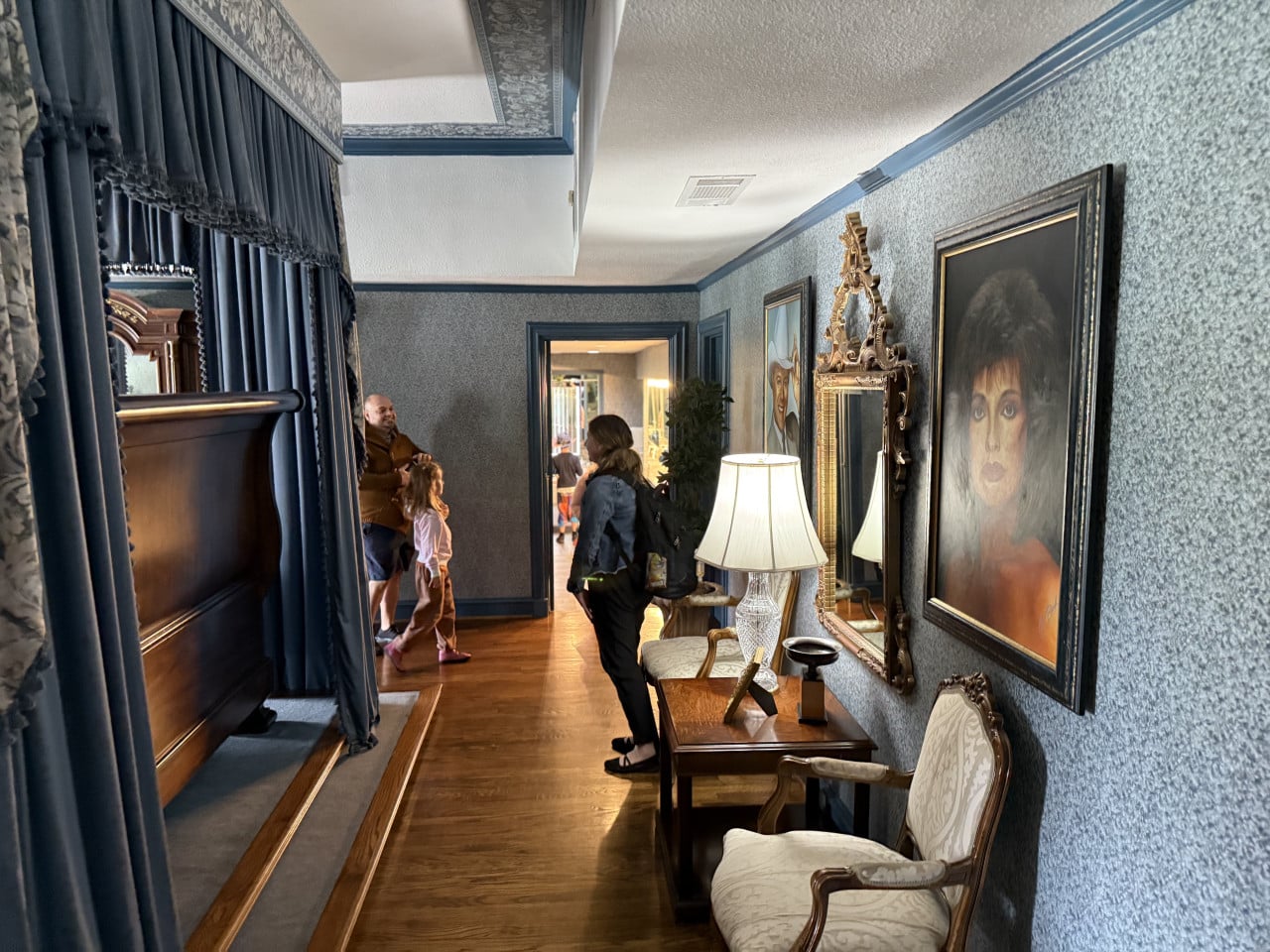 Interiér Southfork Ranch: modrá chodba s lampou, zrcadlem, portrétem a návštěvníky.