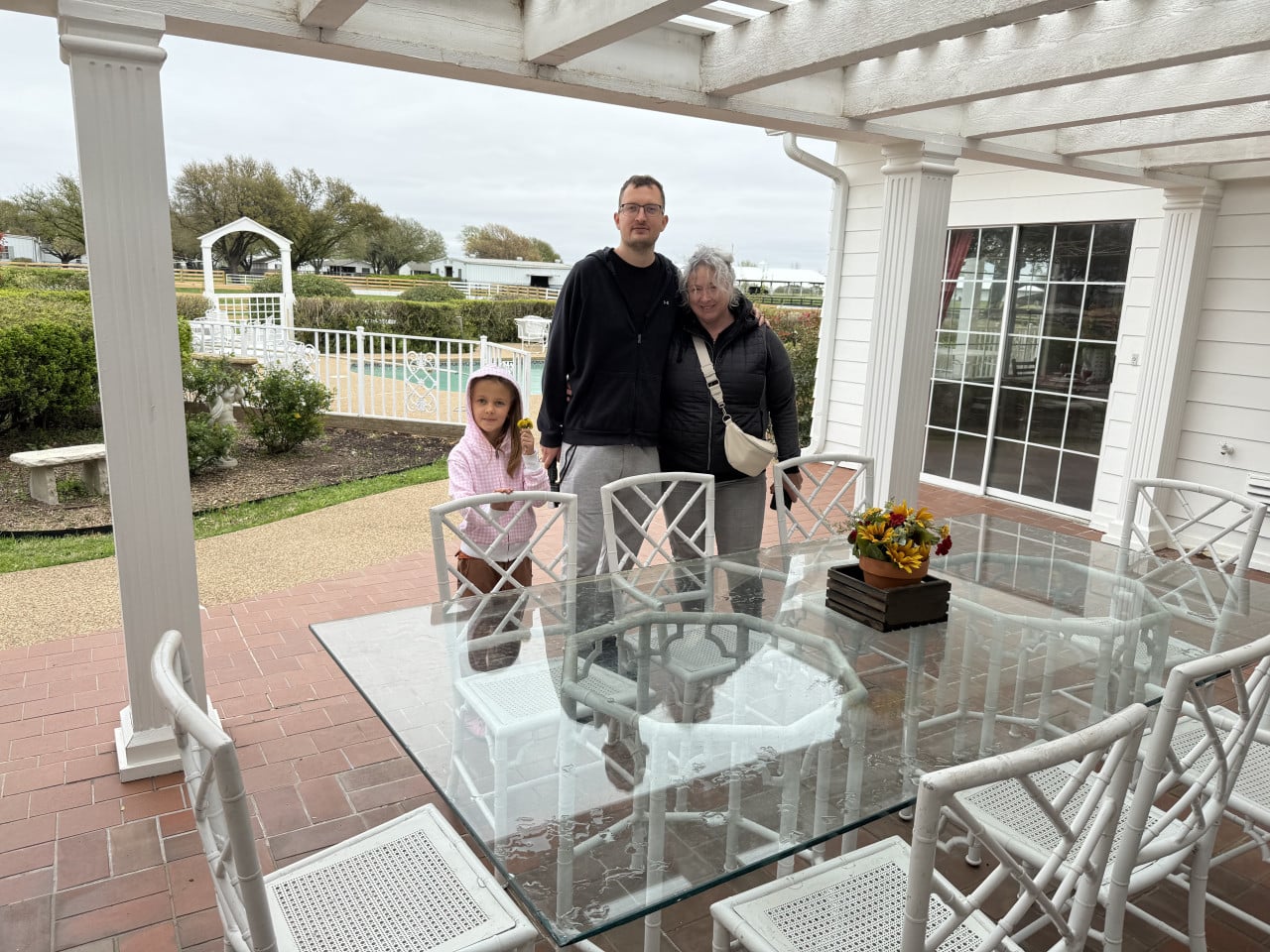 Muž, žena a dítě pózují na verandě u skleněného stolu a bazénu.