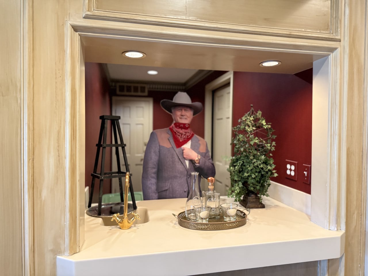 Kartónová postava kovboje u barového pultu s karafou, sklenkami a rostlinou