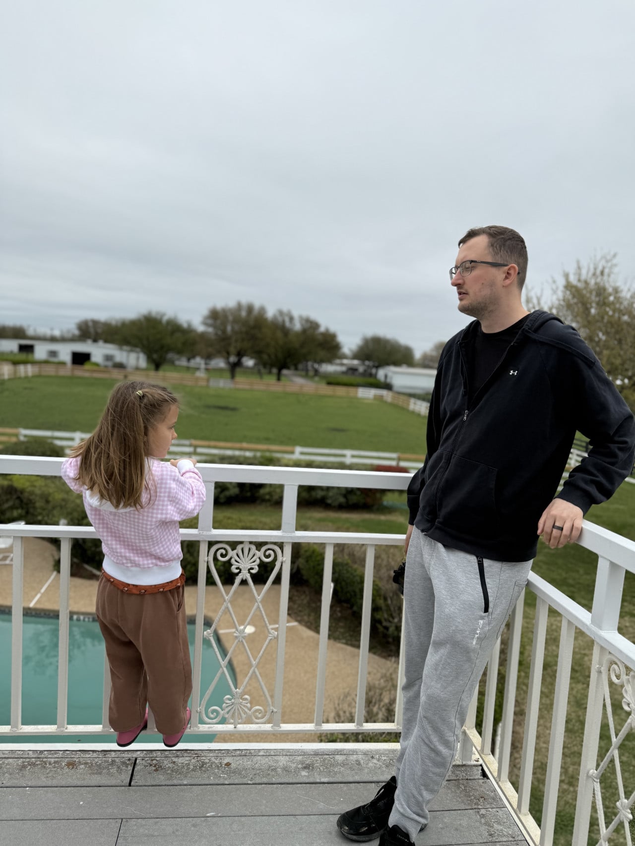 Muž a holčička stojí na balkonu, hledí na zelené pastviny ranče.