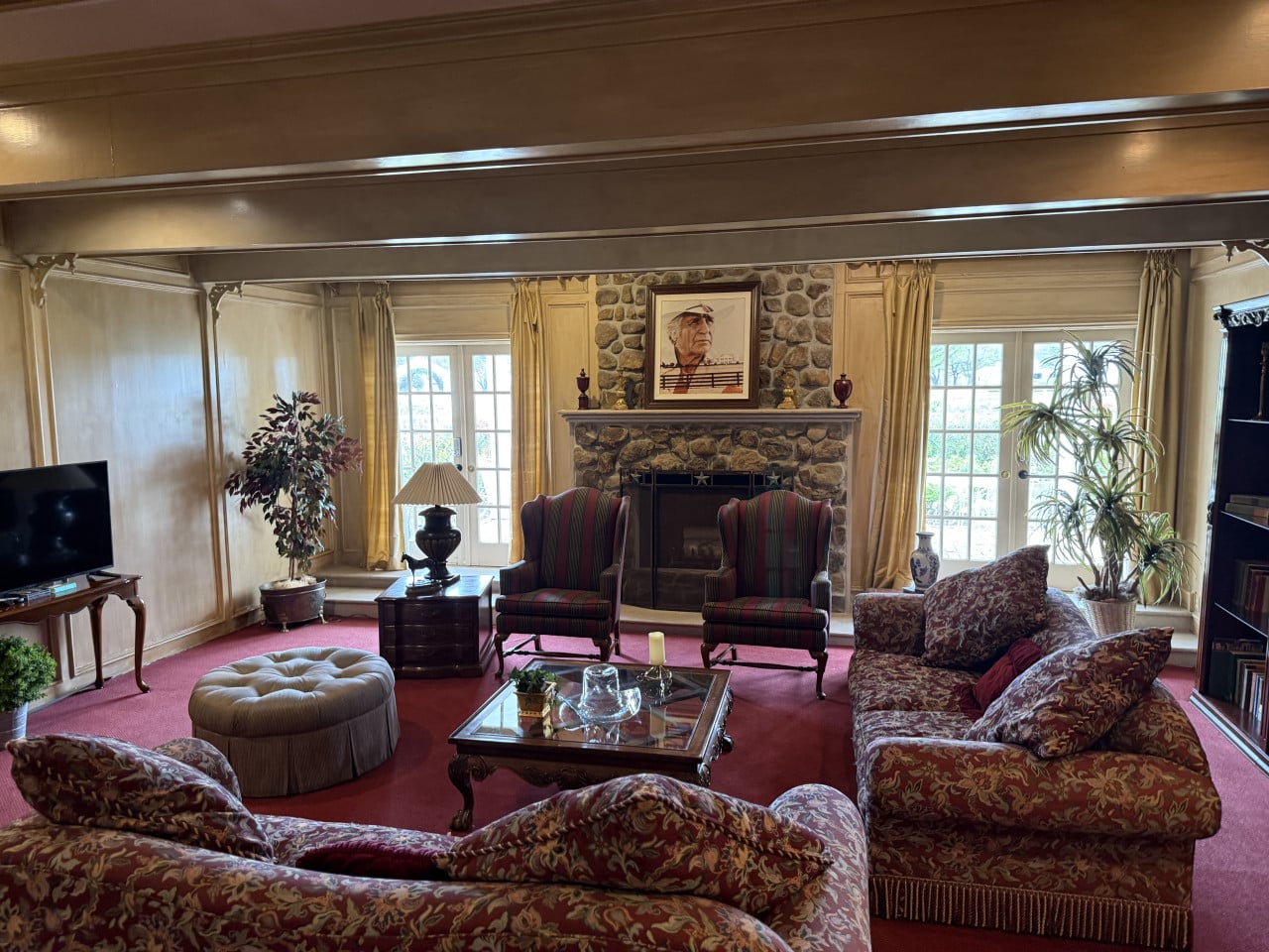 Obývací pokoj Southfork Ranch s krbem z kamene, portrétem, sedačkami a červeným kobercem