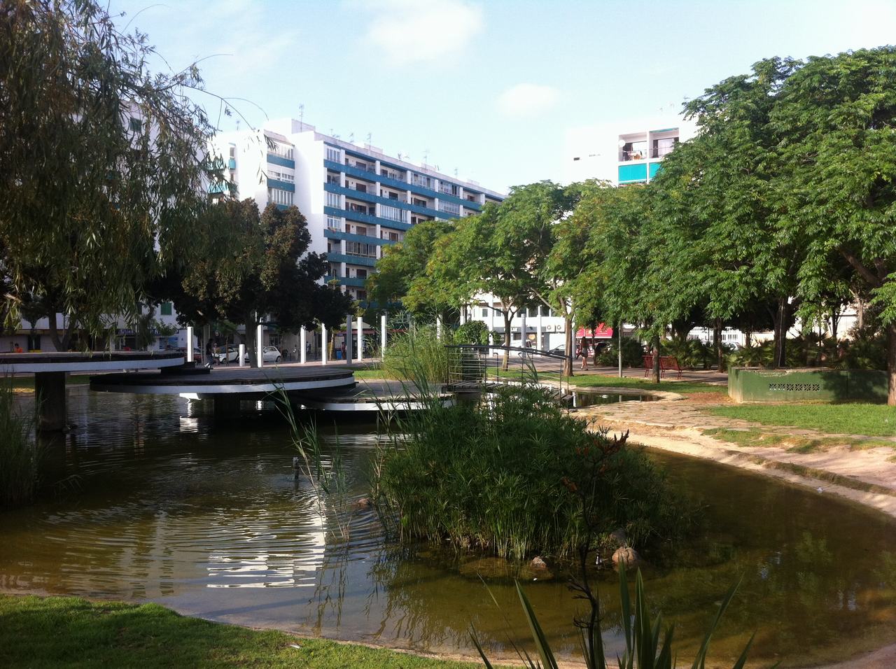 Park s jezírkem a lávkou, stromy a panelové domy v pozadí.