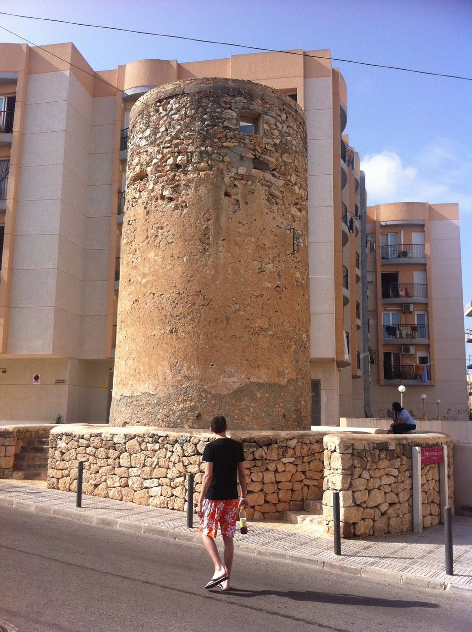 Muž v kraťasech jde kolem kamenné věže před bytovkami, druhý sedí