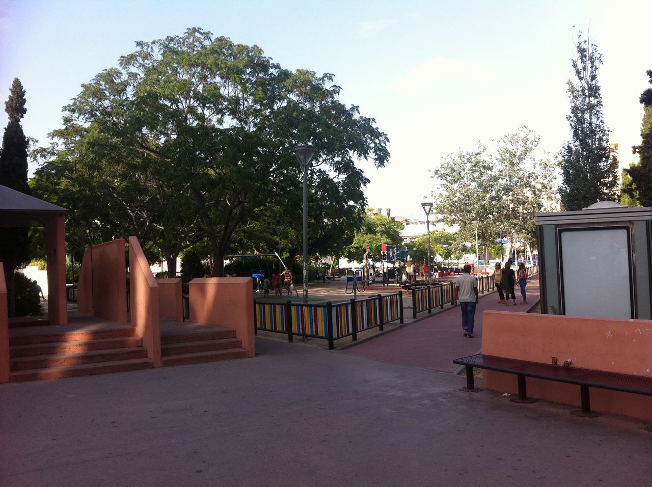 Městský park s dětským hřištěm, barevným plotem, stromy a lidmi na chodníku