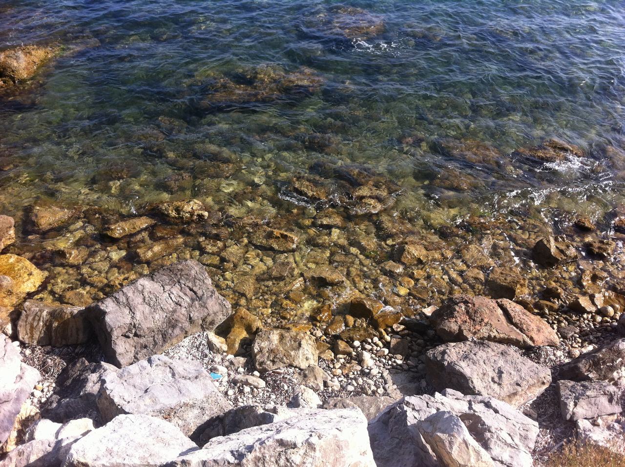 Průzračná mořská voda omývá kamenité pobřeží s velkými balvany na slunci.