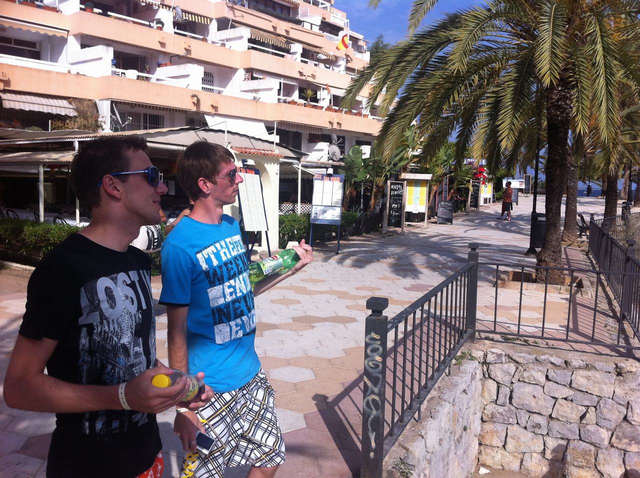 Dva muži v brýlích s lahvemi na promenádě u palem a hotelů