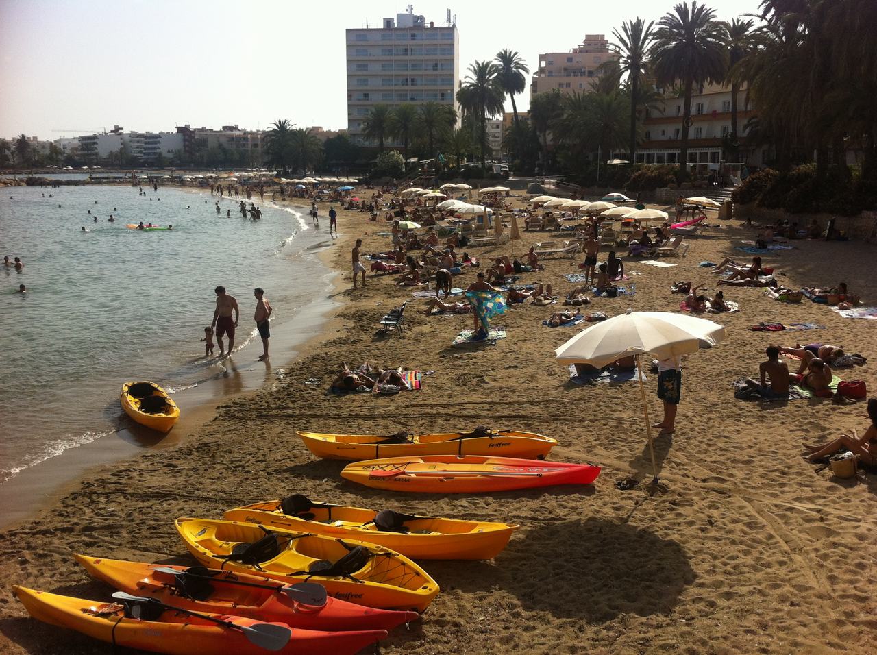 Plná pláž s koupajícími se lidmi, slunečníky a kajaky na písku