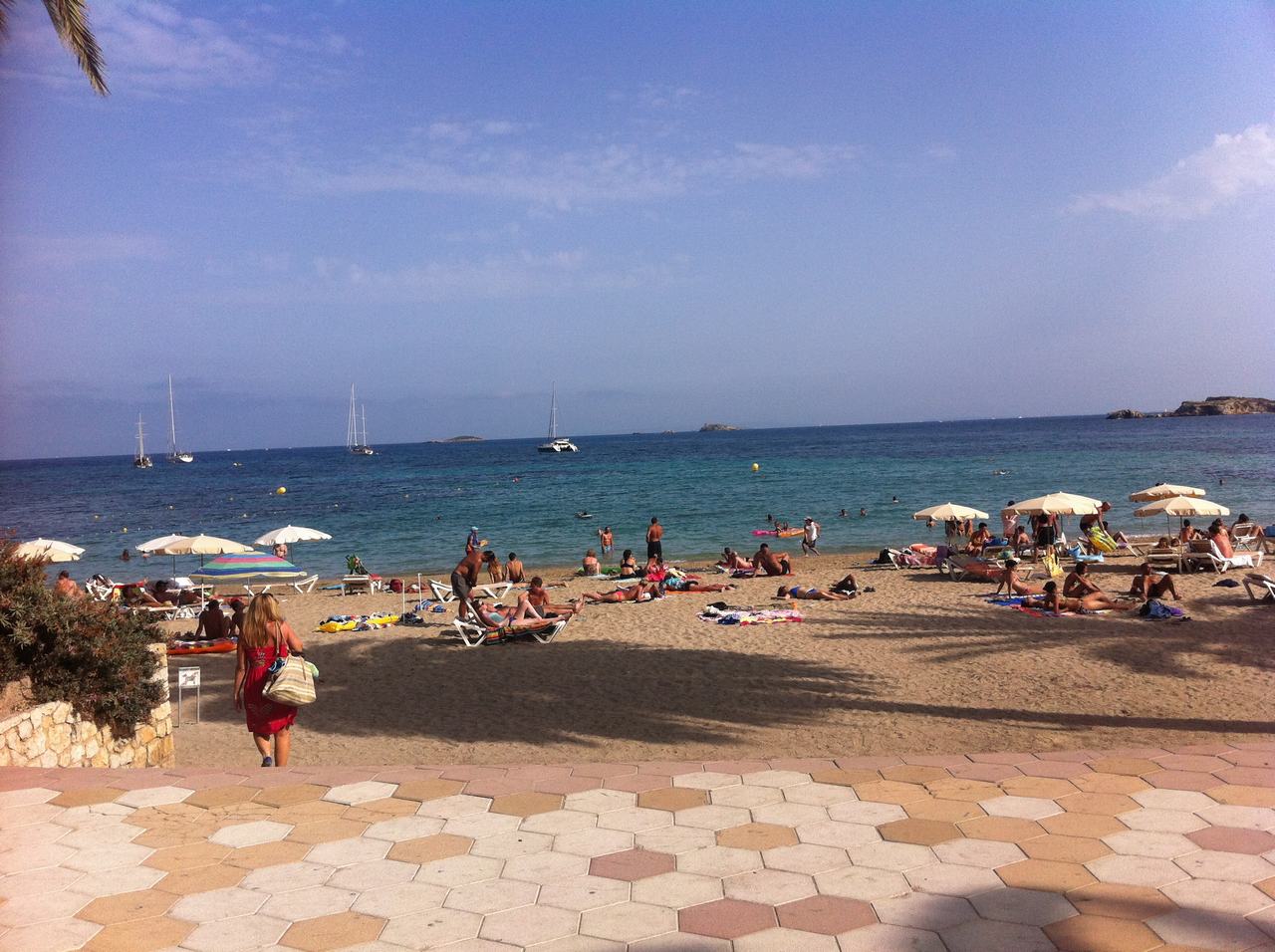 Písečná pláž s lidmi na lehátkách, slunečníky, moře a plachetnice v dálce