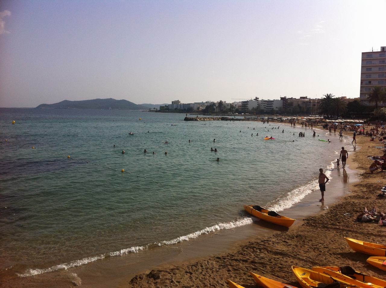 Písečná pláž s lidmi ve vodě, žluté kajaky na břehu, město v pozadí