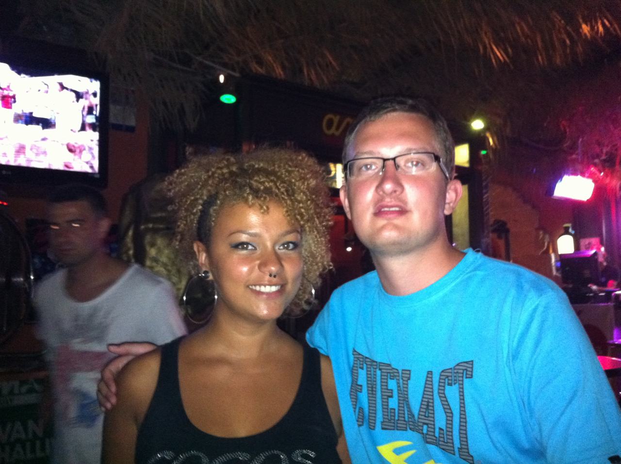 Dva lidé pózují v nočním baru; muž v tričku s nápisem EVERLAST.