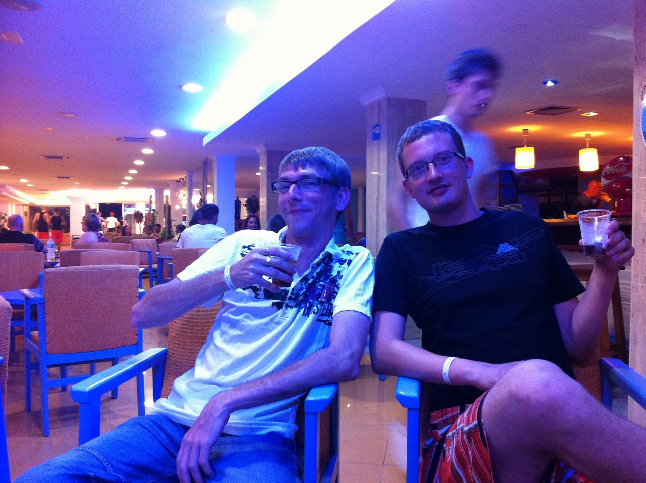 Dva muži sedí v hotelovém baru, drží kelímky; tričko Kappa, logo Coca-Cola.