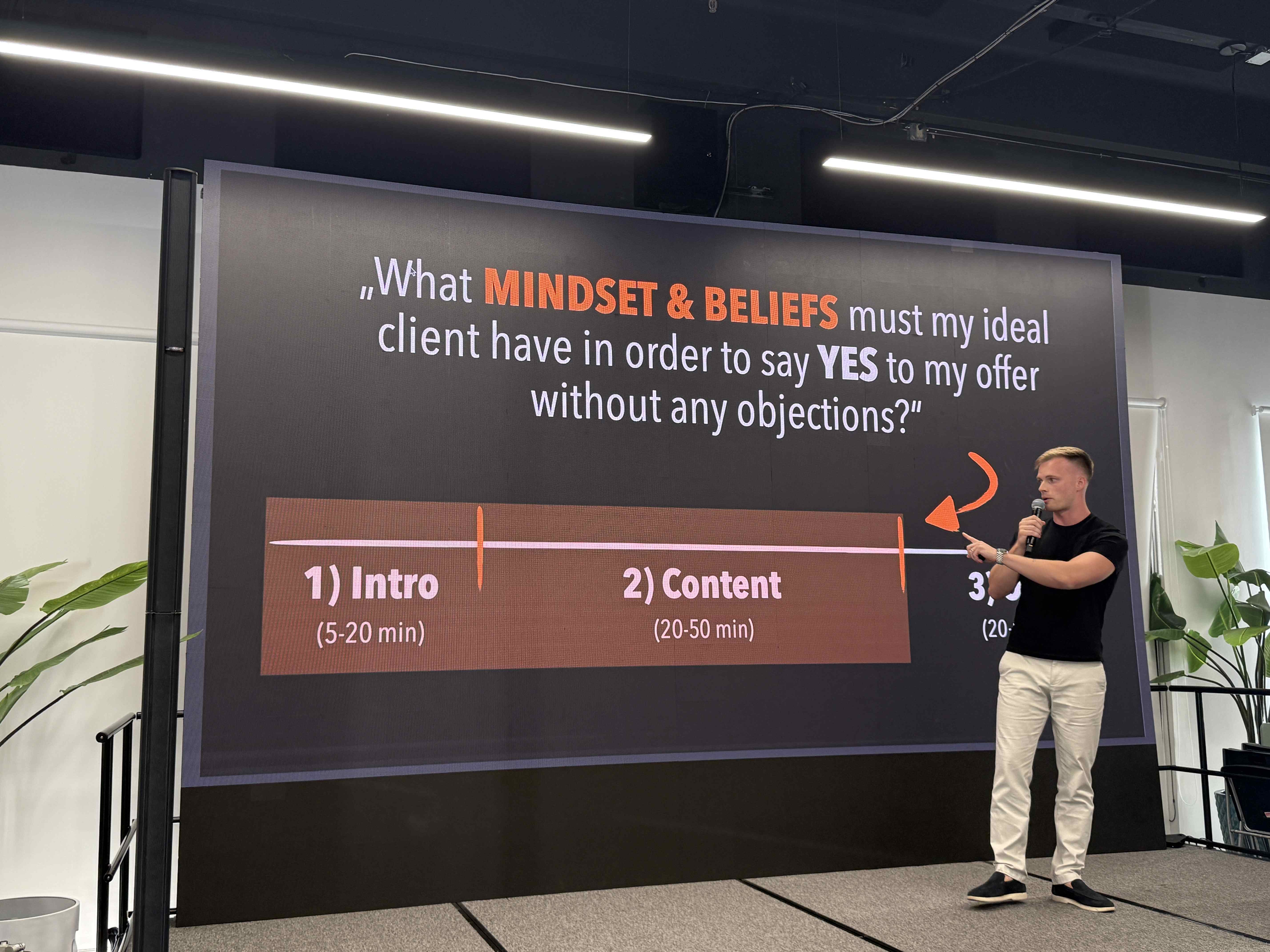 Řečník na pódiu ukazuje slide What MINDSET & BELIEFS; část agendy Intro, Content.