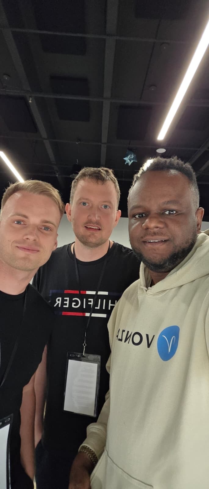 Tři muži na selfie v moderním interiéru, dva mají jmenovky a mikiny s logy značek.