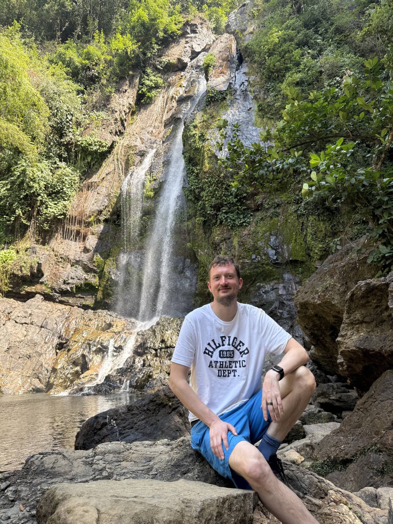 Muž sedí na skále před vodopádem; tričko HILFIGER 1985 ATHLETIC DEPT.
