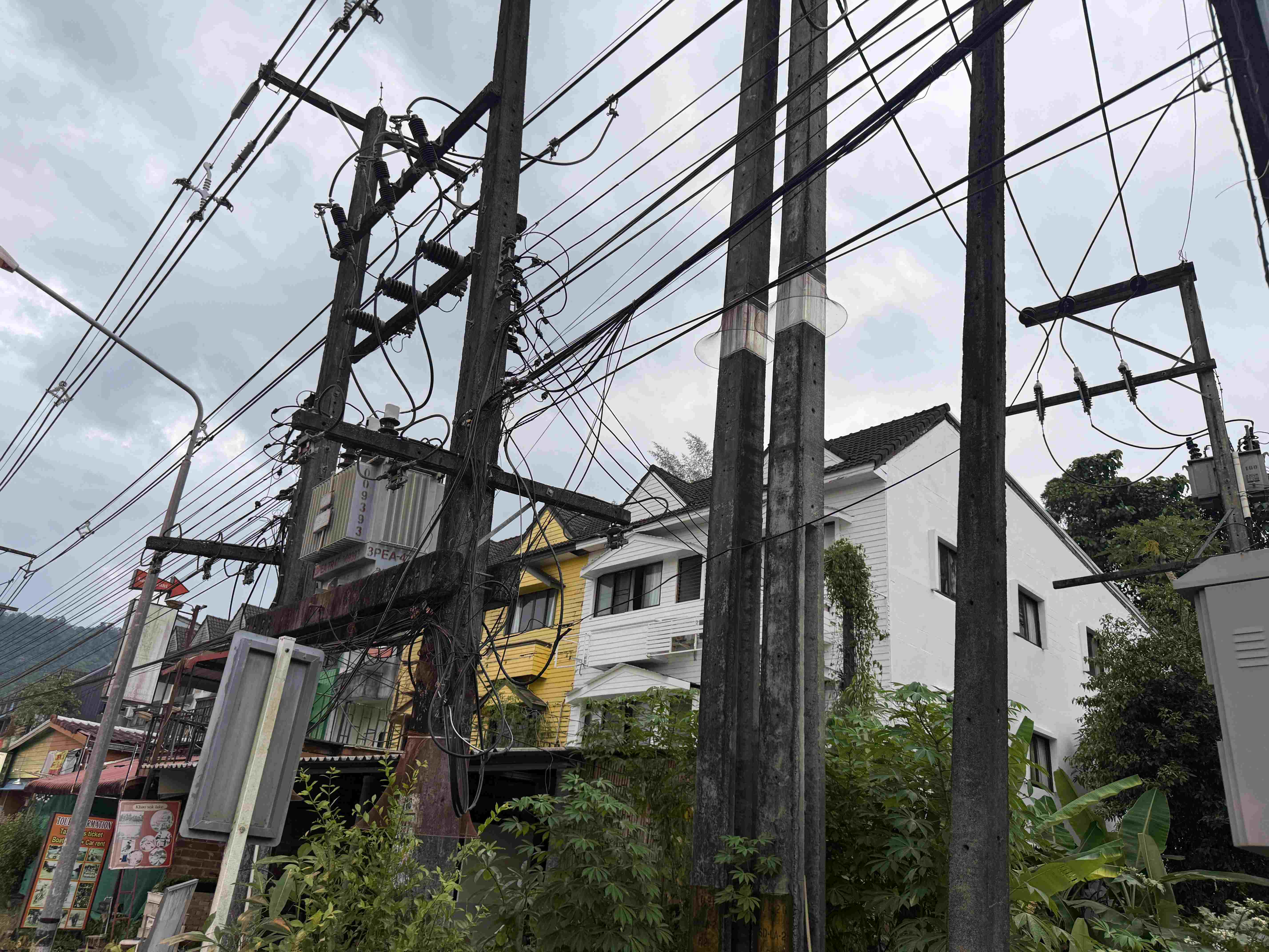 Zamotané elektrické kabely na sloupech nad ulicí, domy v pozadí.