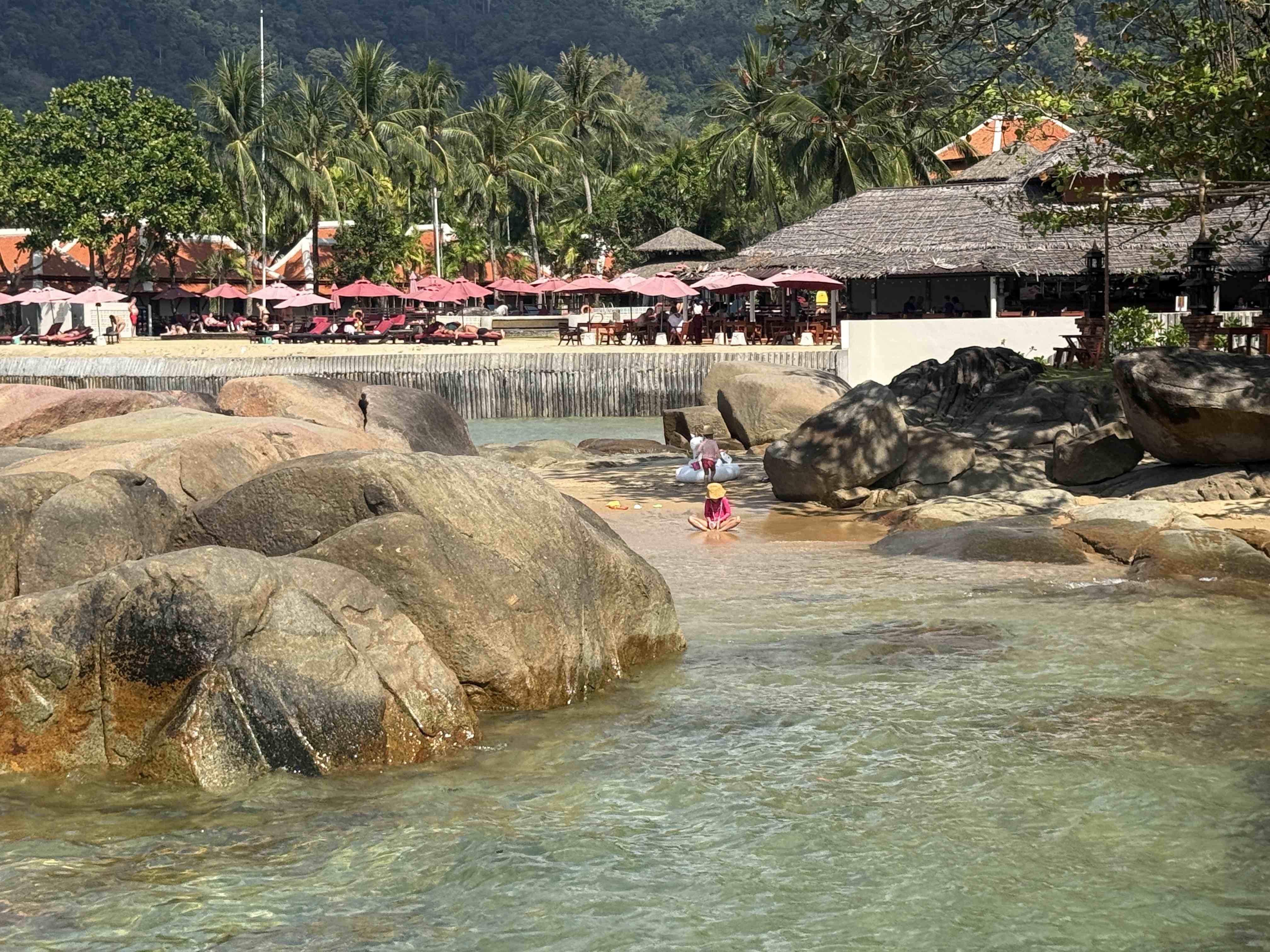 Děti se brodí v mělké laguně mezi balvany, v pozadí plážový resort s růžovými slunečníky