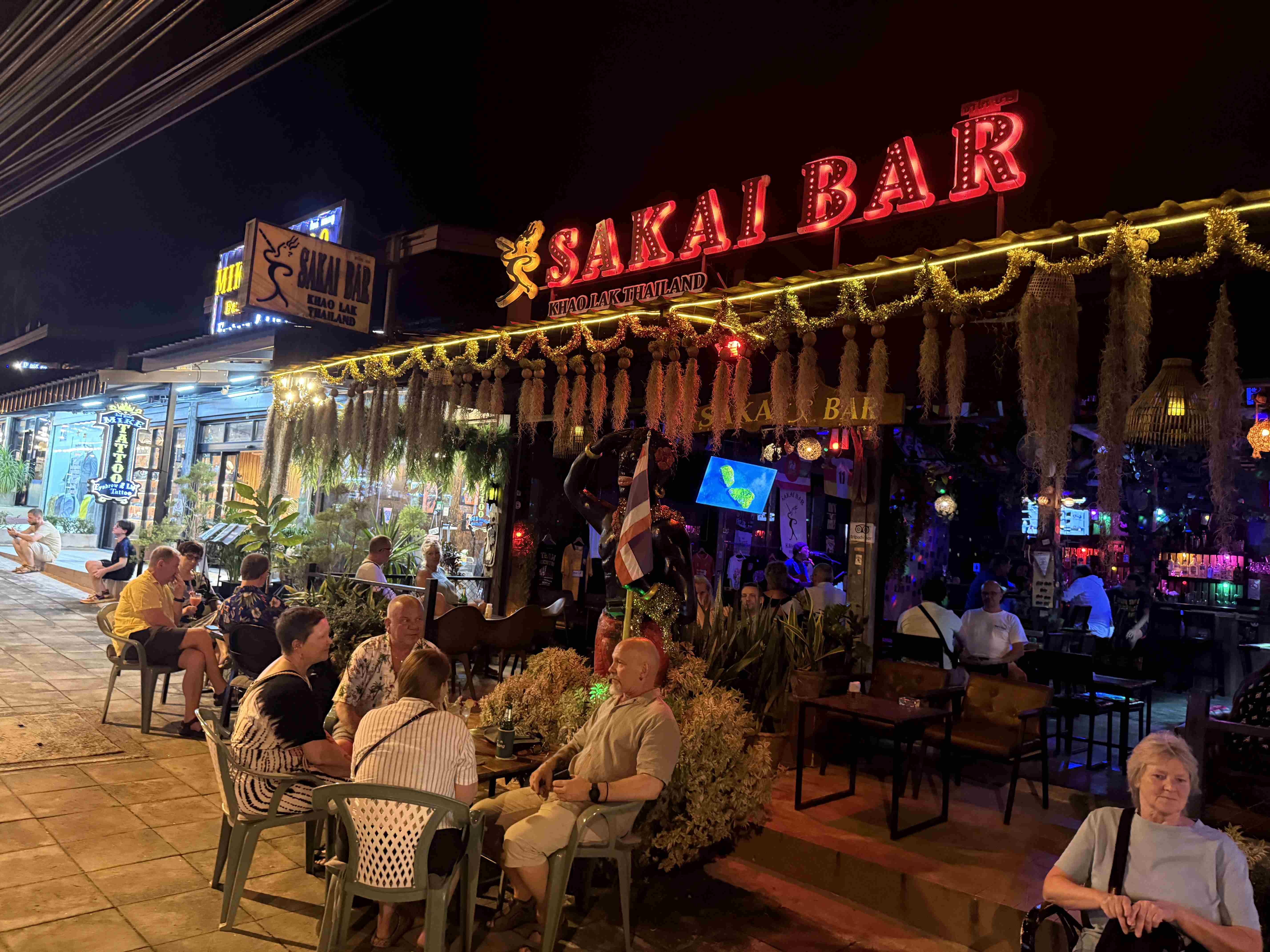 Noční terasa s hosty pod neonem SAKAI BAR, KHAO LAK THAILAND.