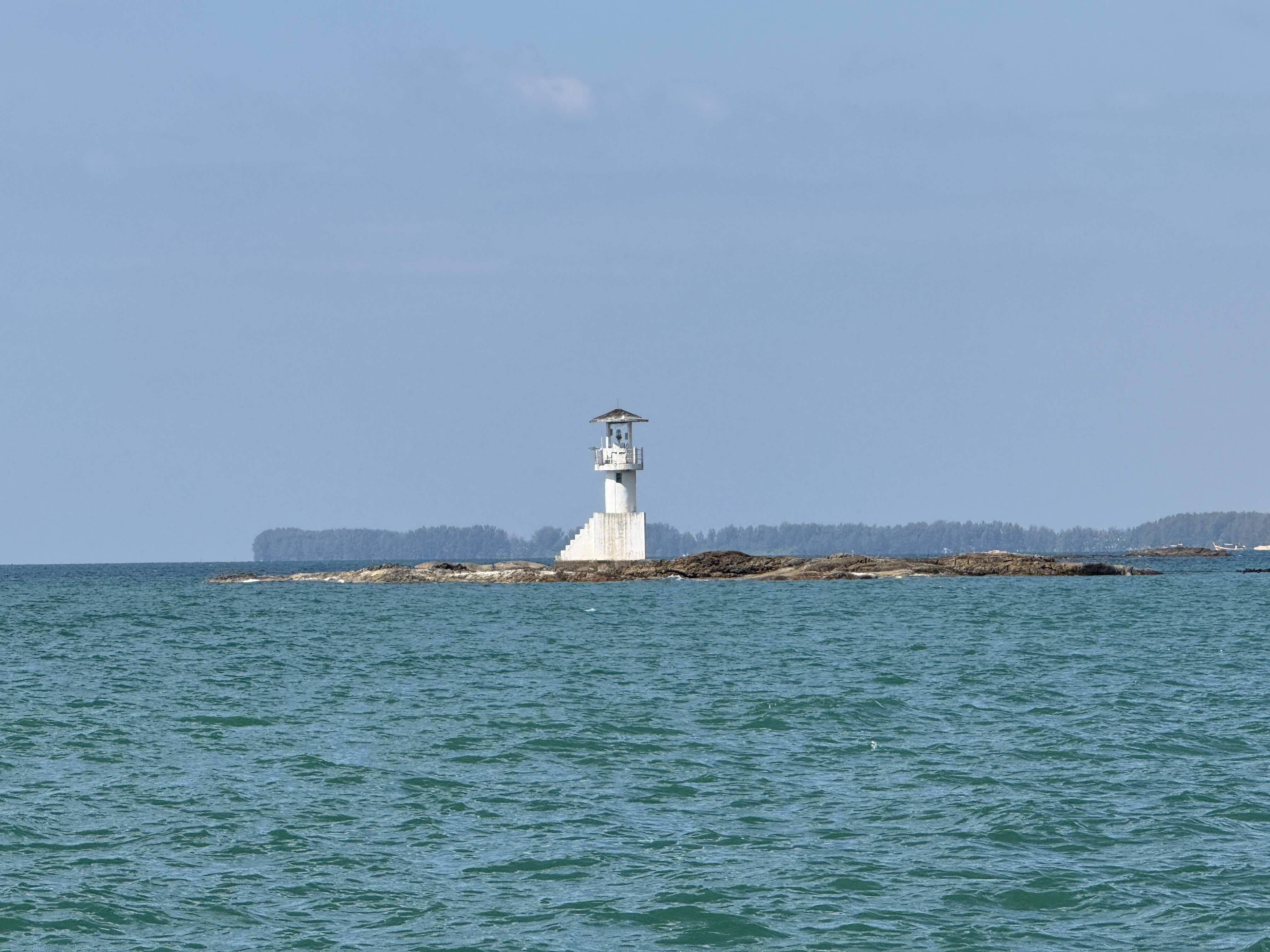 Bílý maják na skalnatém ostrůvku uprostřed tyrkysového moře, v dálce pobřeží