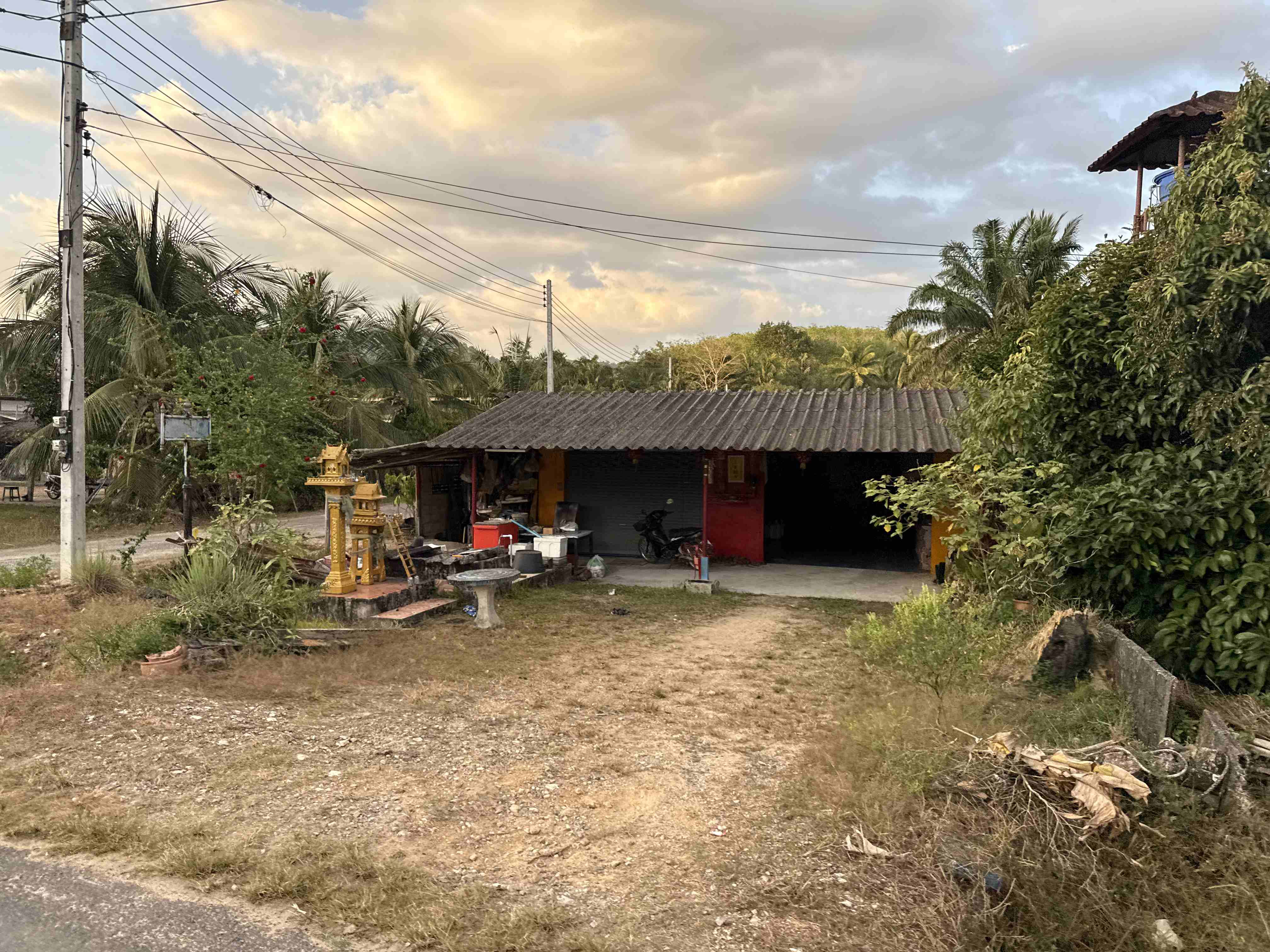 Venkovský dům s plechovou střechou, thajský spirit house, palmy a elektrické vedení.