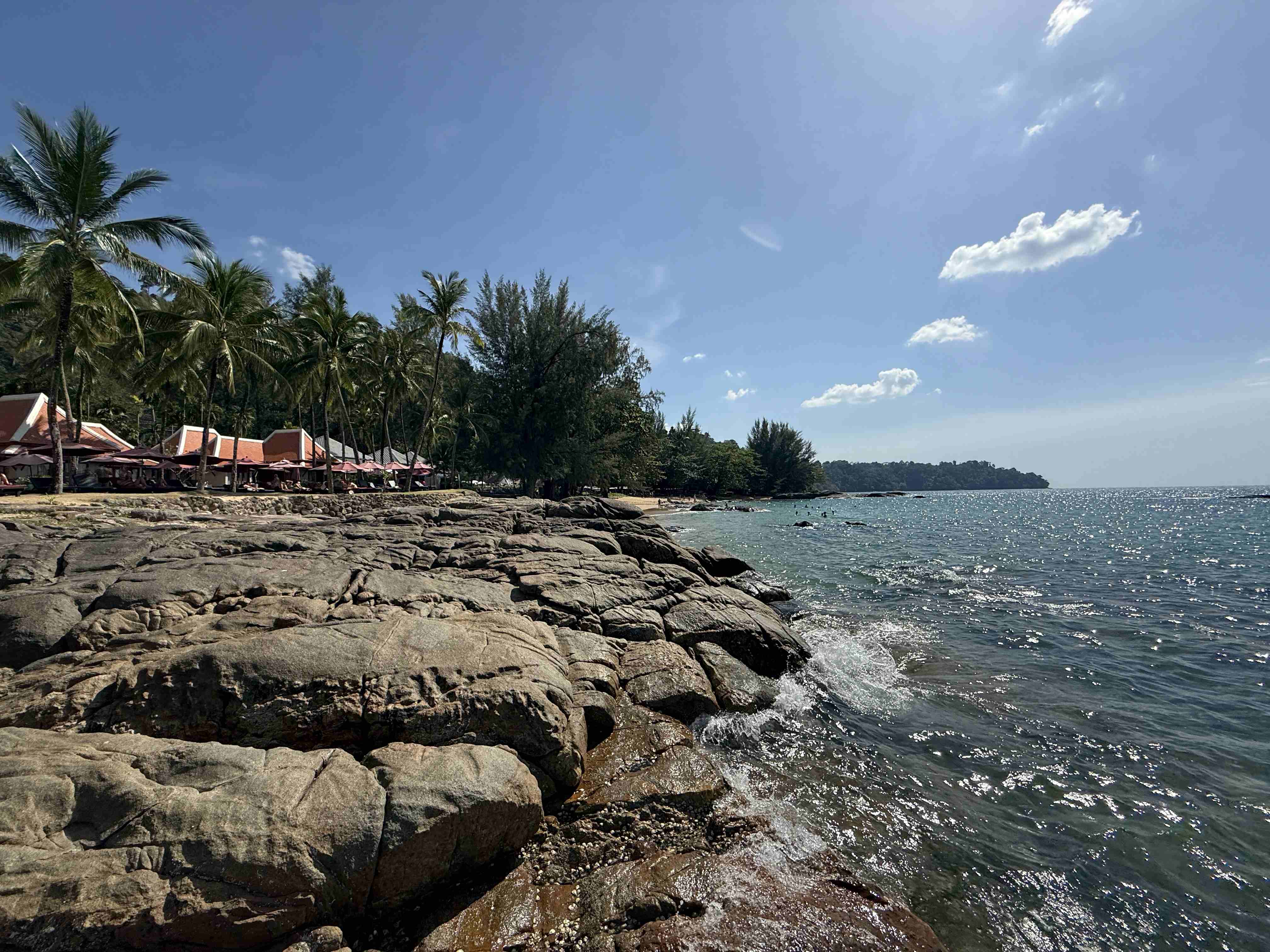 Skalnaté pobřeží u moře, palmy a resortové bungalovy pod modrou oblohou.