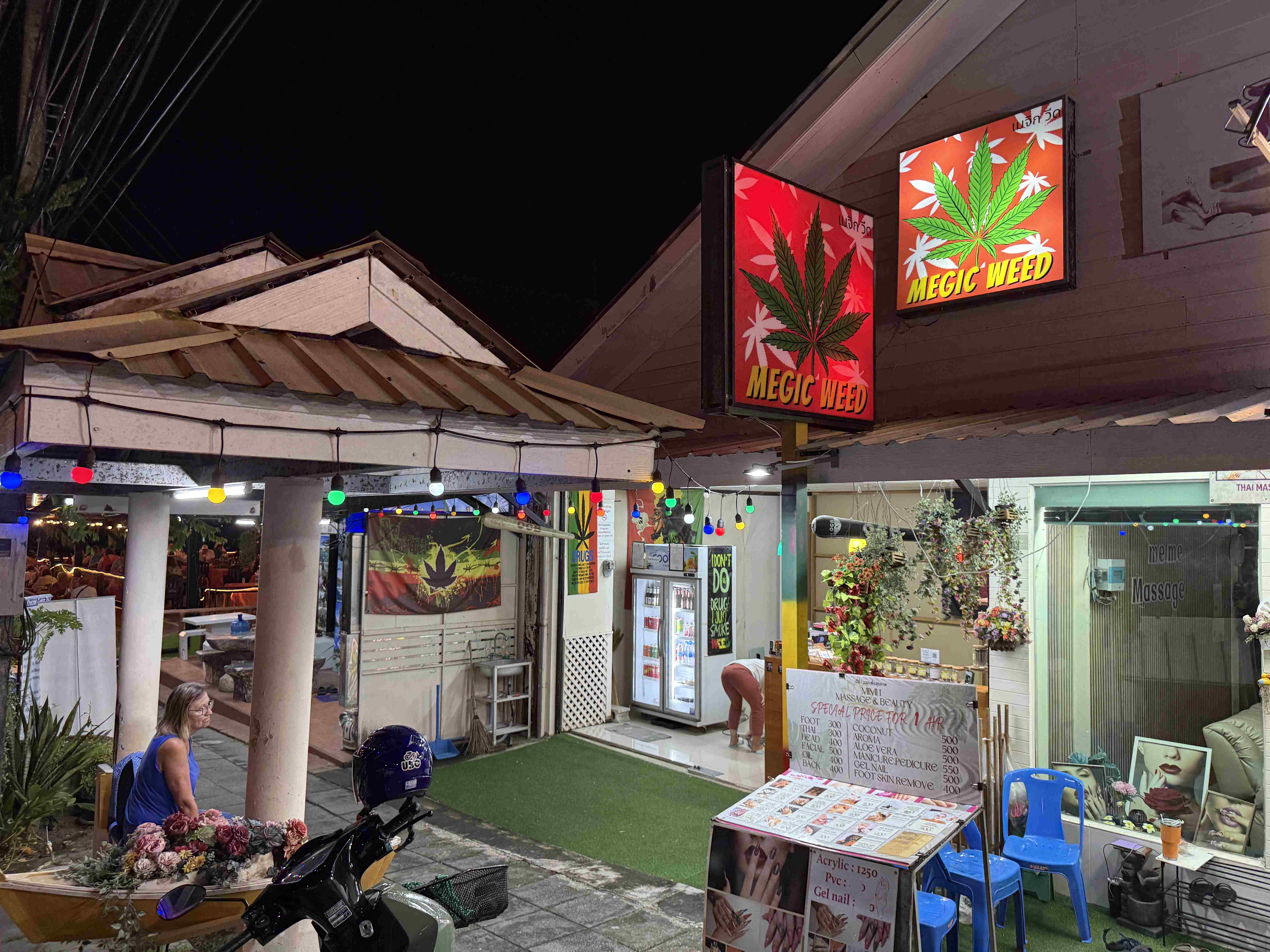 Noční ulice s osvětlenými cedulemi MEGIC WEED a thajskými masážemi, skútrem