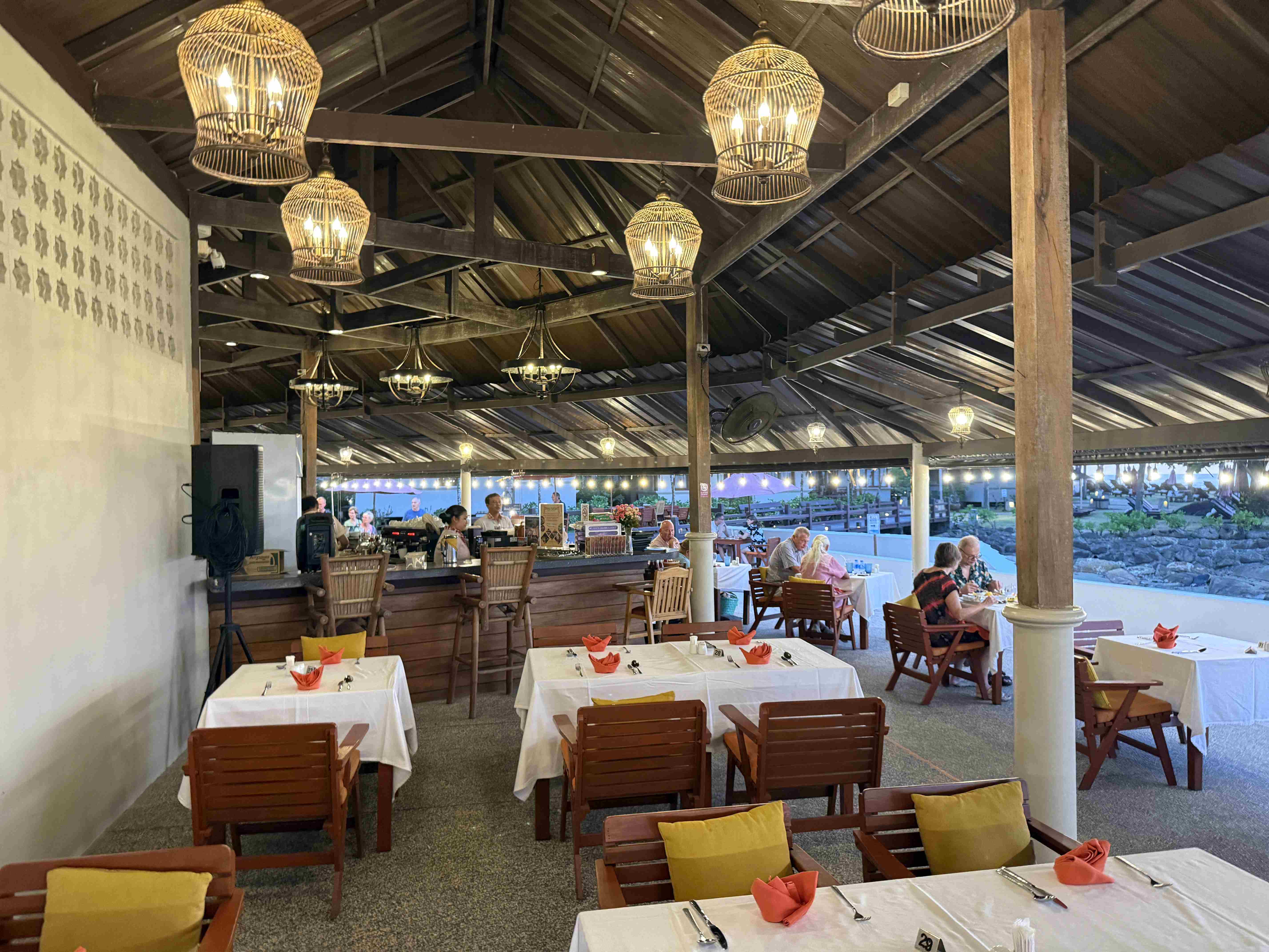 Otevřená hotelová restaurace s barem, dřevěným stropem, lampami a prostřenými stoly.