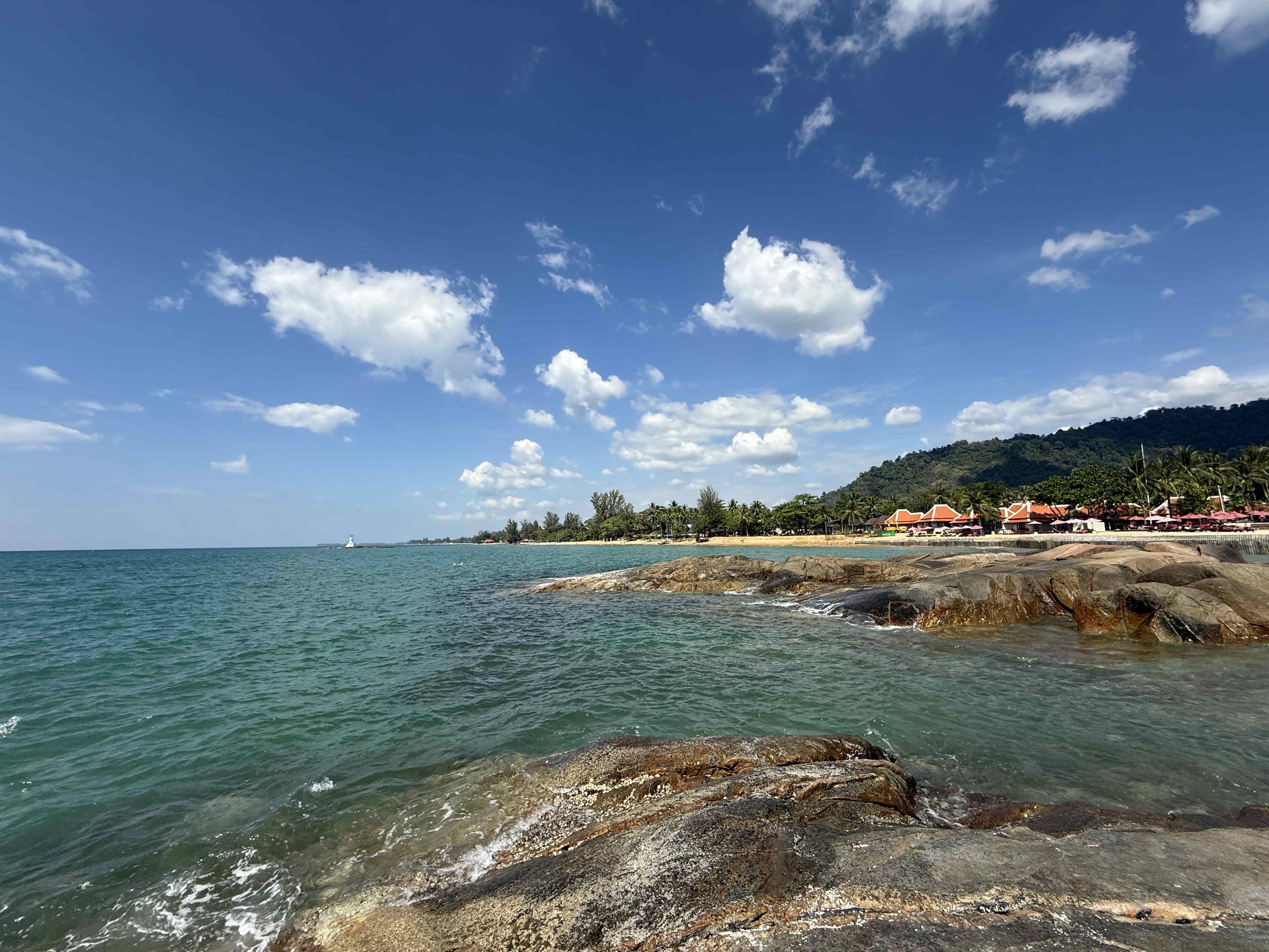 Skalnaté pobřeží a tyrkysové moře u pláže Khao Lak, v dálce resort